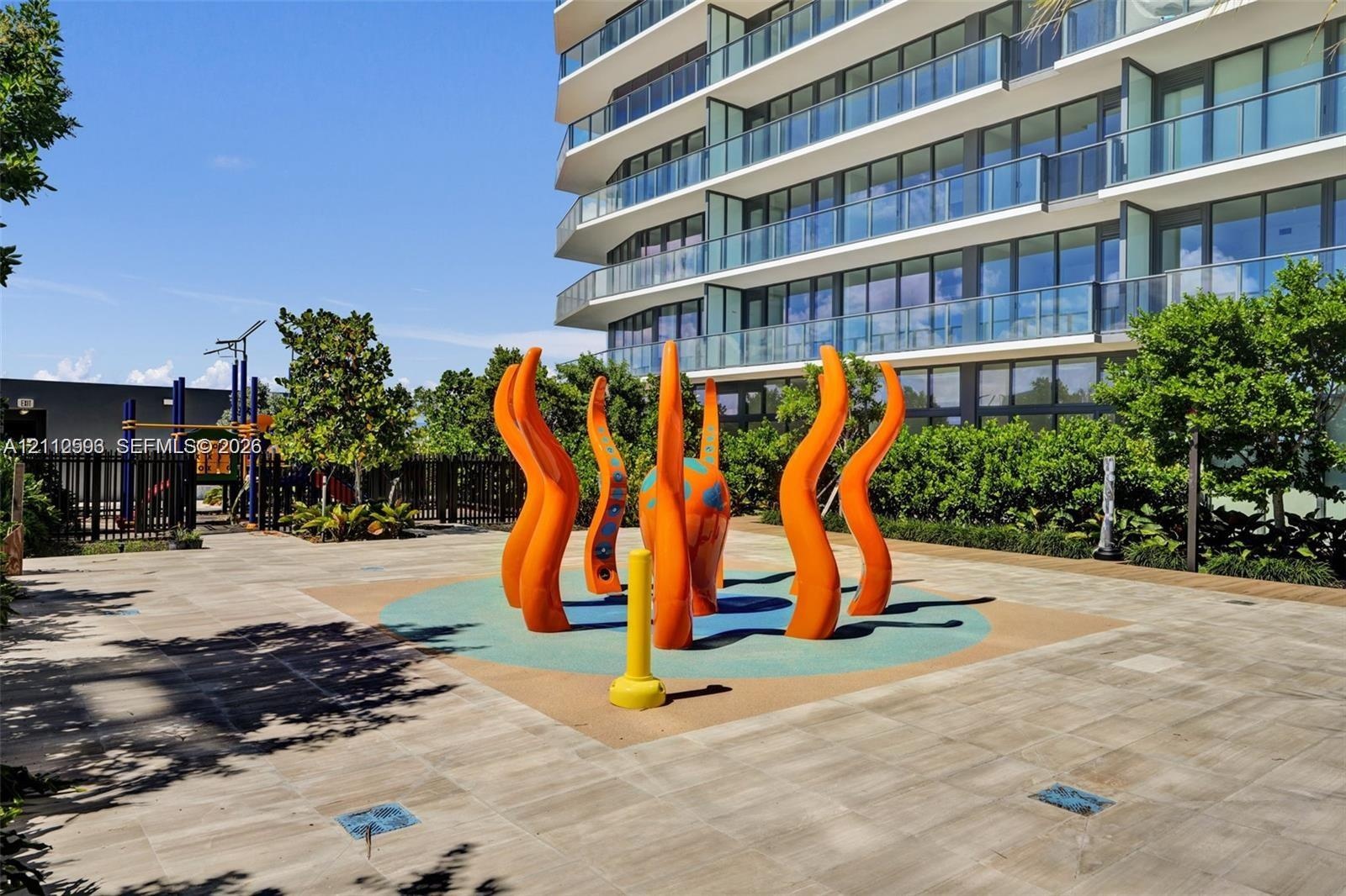 Outdoor recreation area featuring a paved terrace, colorful sculptural water feature, integrated rubberized ground surface, and perimeter landscaping
