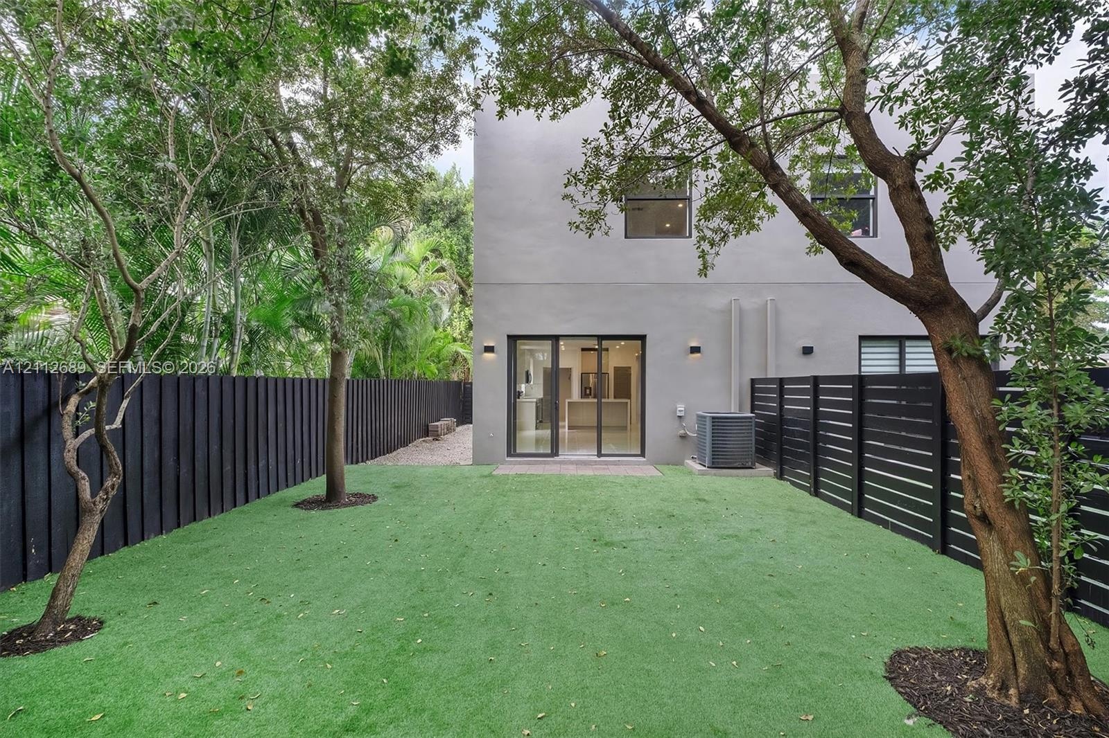 Fenced backyard featuring synthetic turf, mature trees, smooth stucco exterior, and sliding glass doors