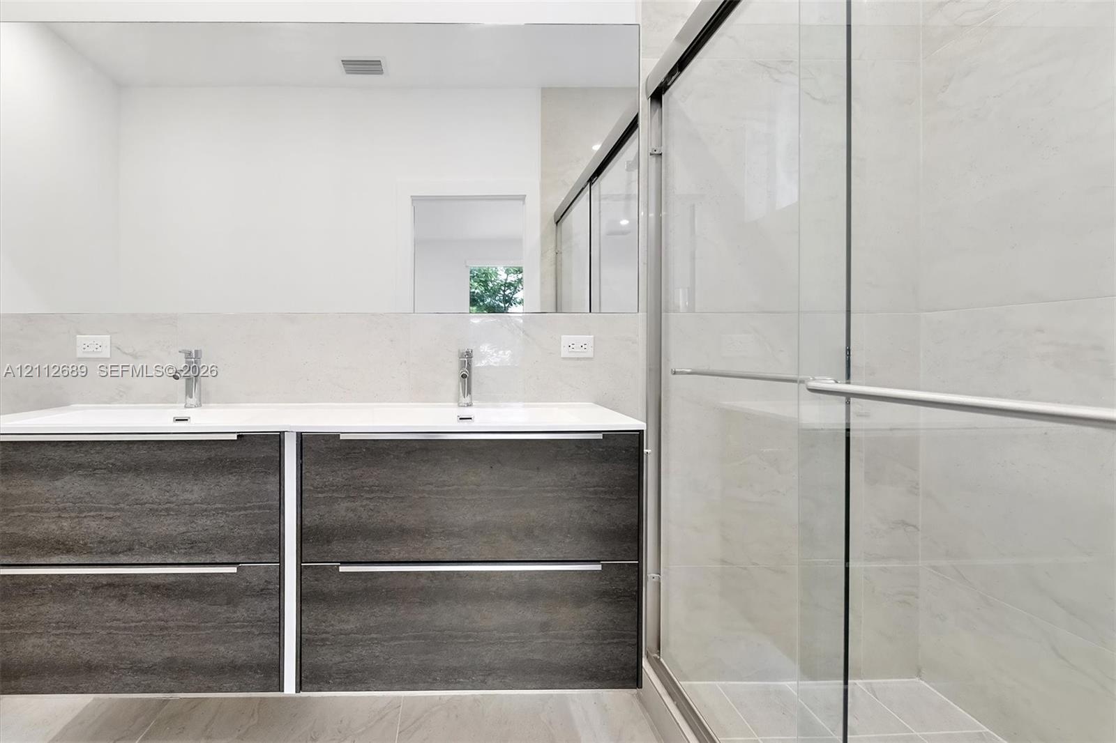 Floating wood-finish double vanity with dual undermount sinks and chrome fixtures, large wall-mounted mirror, and walk-in shower featuring glass slidi