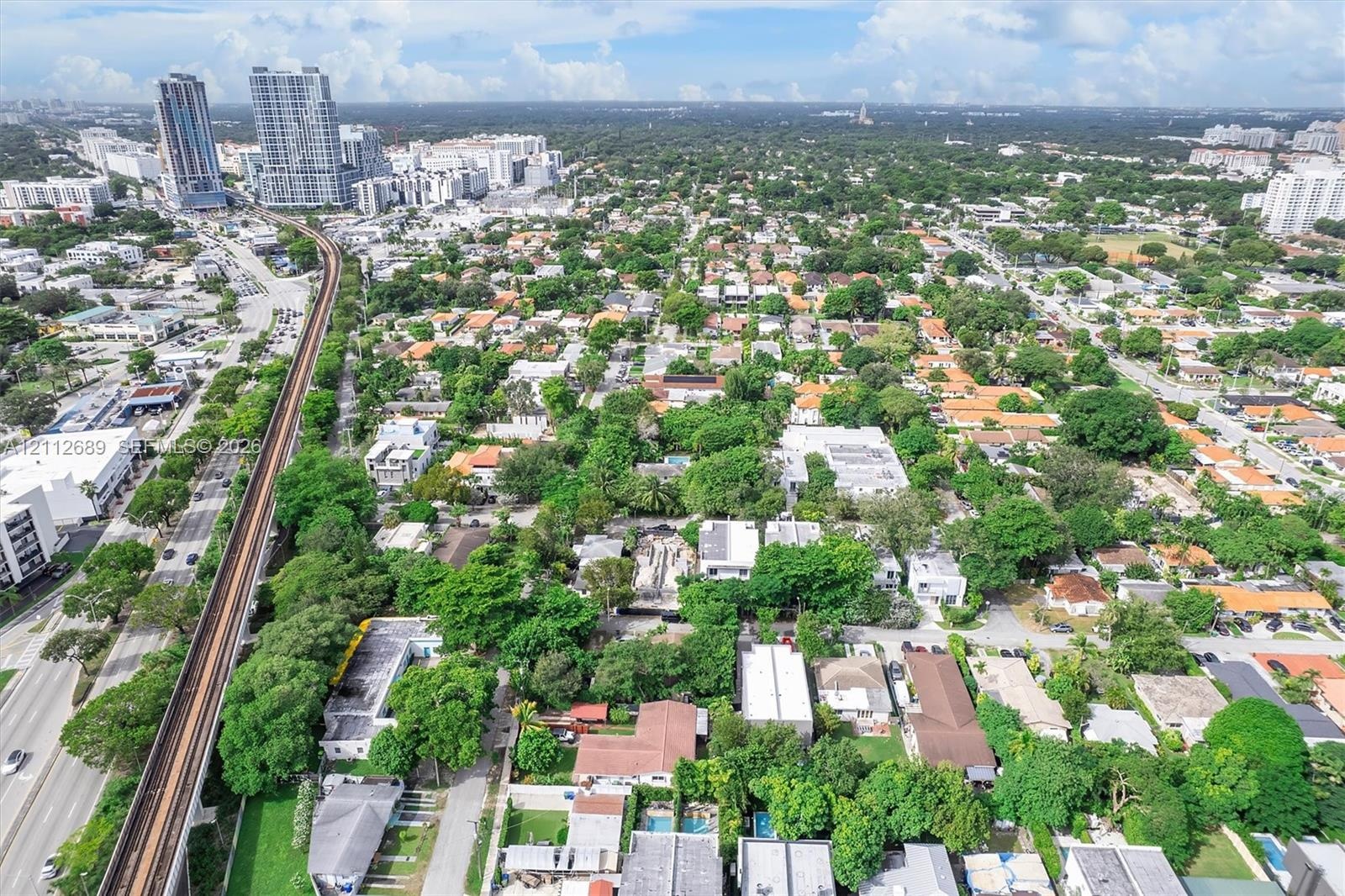 Aerial perspective of an urban setting featuring high-rise buildings, a nearby rail line, a dense canopy of mature trees, and established residential 