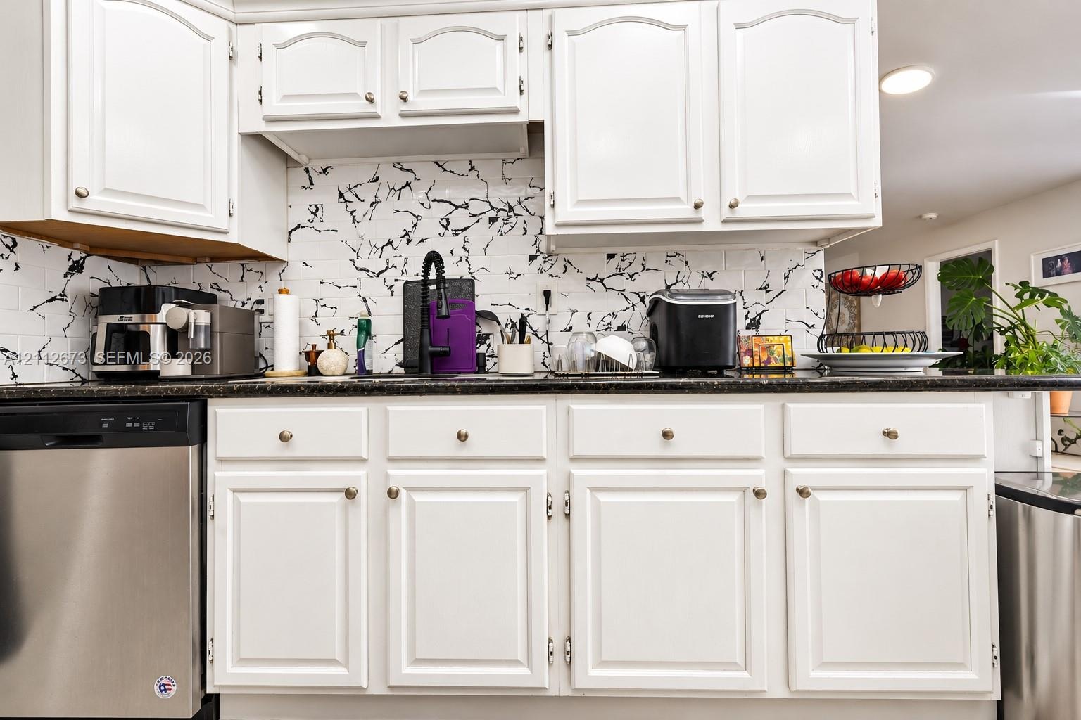 White raised-panel cabinetry, dark stone-finish countertops, high-contrast patterned backsplash, and stainless steel dishwasher
