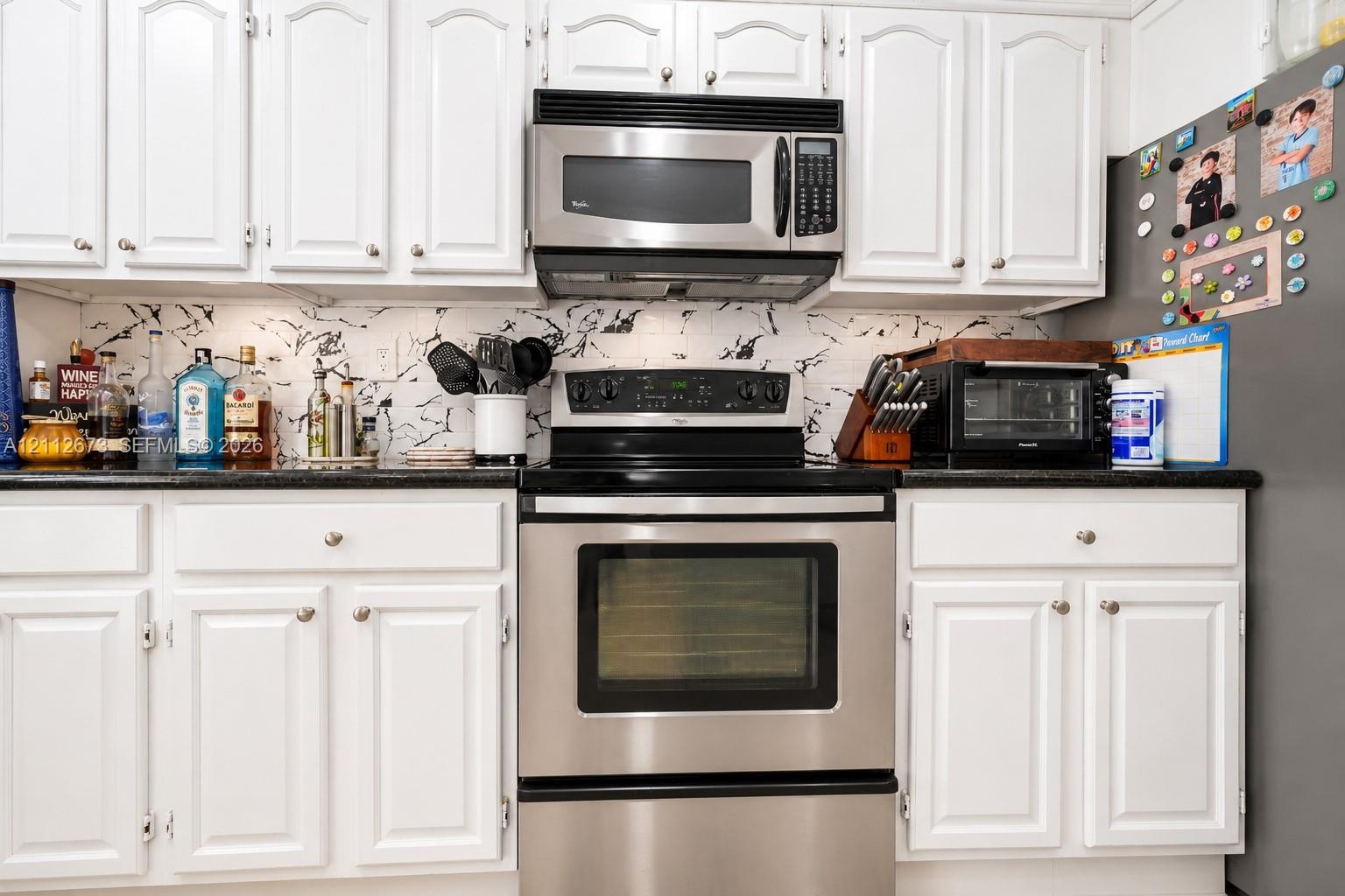 Kitchen featuring white raised-panel cabinetry, dark solid-surface countertops, patterned tile backsplash, and stainless steel appliances including an