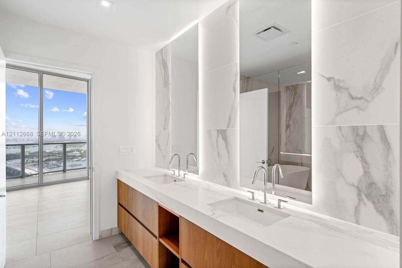 Dual-sink vanity with wood-finish cabinetry, stone-surface countertop, floor-to-ceiling stone-tiled backsplash with integrated vertical lighting, and 