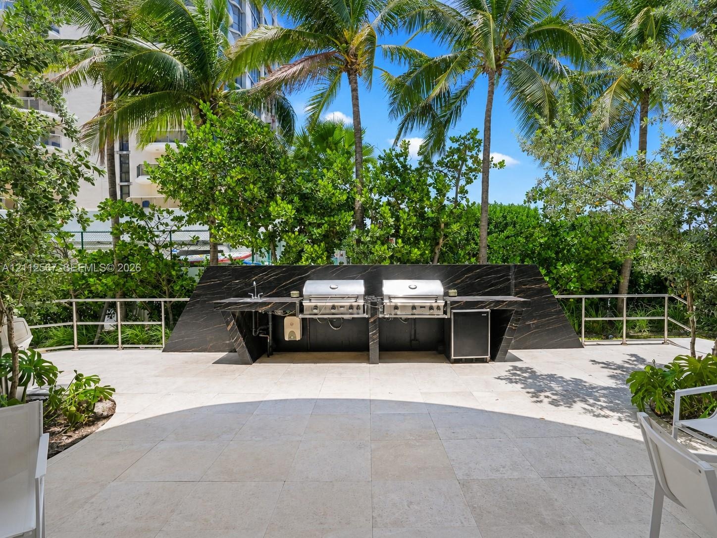Outdoor kitchen featuring a dark stone-finish backsplash and countertop, stainless steel dual grills, integrated refrigeration, and a stone-tiled pati