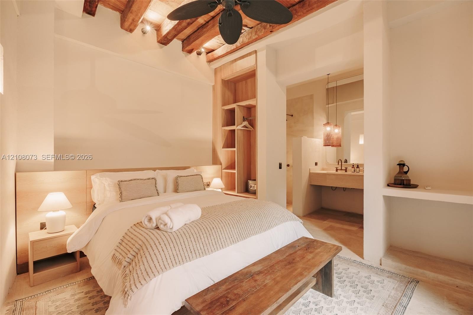 Exposed timber ceiling beams, built-in floor-to-ceiling wooden shelving, integrated wall-mounted headboard, and floating bathroom vanity with an under