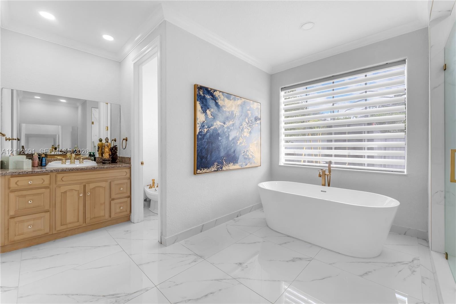 Freestanding soaking tub with gold-tone fixtures, wood cabinetry with stone-surface countertop, white tile flooring with veining detail, and large-for