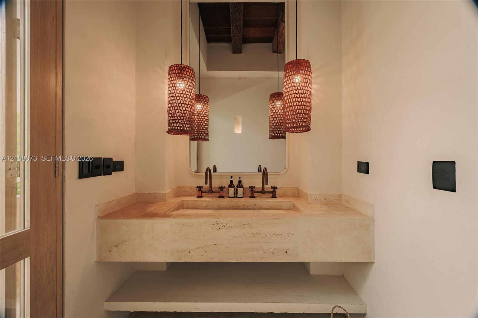 Custom stone floating vanity with an integrated basin, dual wall-mounted faucets, arched wall mirror, and exposed timber ceiling beams