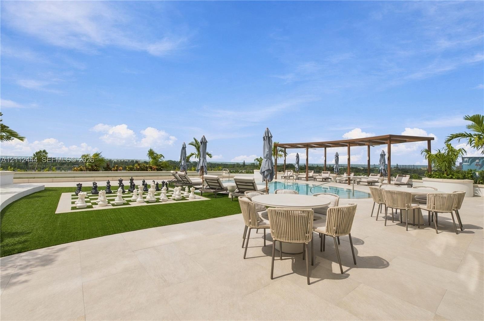 Outdoor terrace featuring light-toned stone paving, a swimming pool, a wood-frame pergola, synthetic turf, and an oversized permanent outdoor chess bo