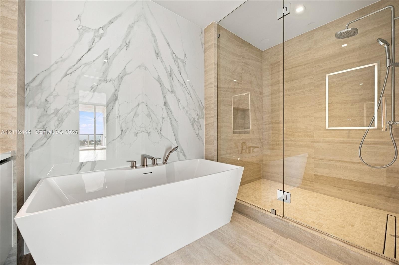 Freestanding soaking tub with a wall-mounted faucet, large-format stone-tile wall, glass-enclosed shower, integrated rainfall showerhead, and recessed
