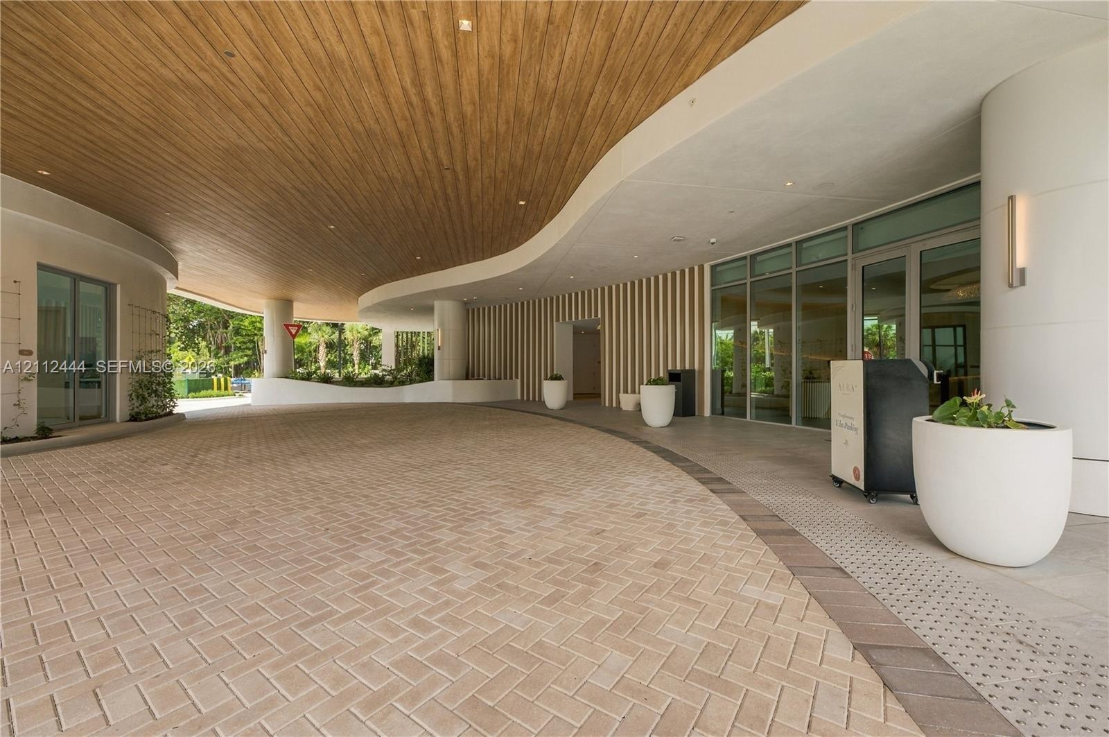 Covered porte-cochère featuring a curved wood-panel ceiling, herringbone-pattern paver drive, vertical slat wall detailing, and expansive glass entry 