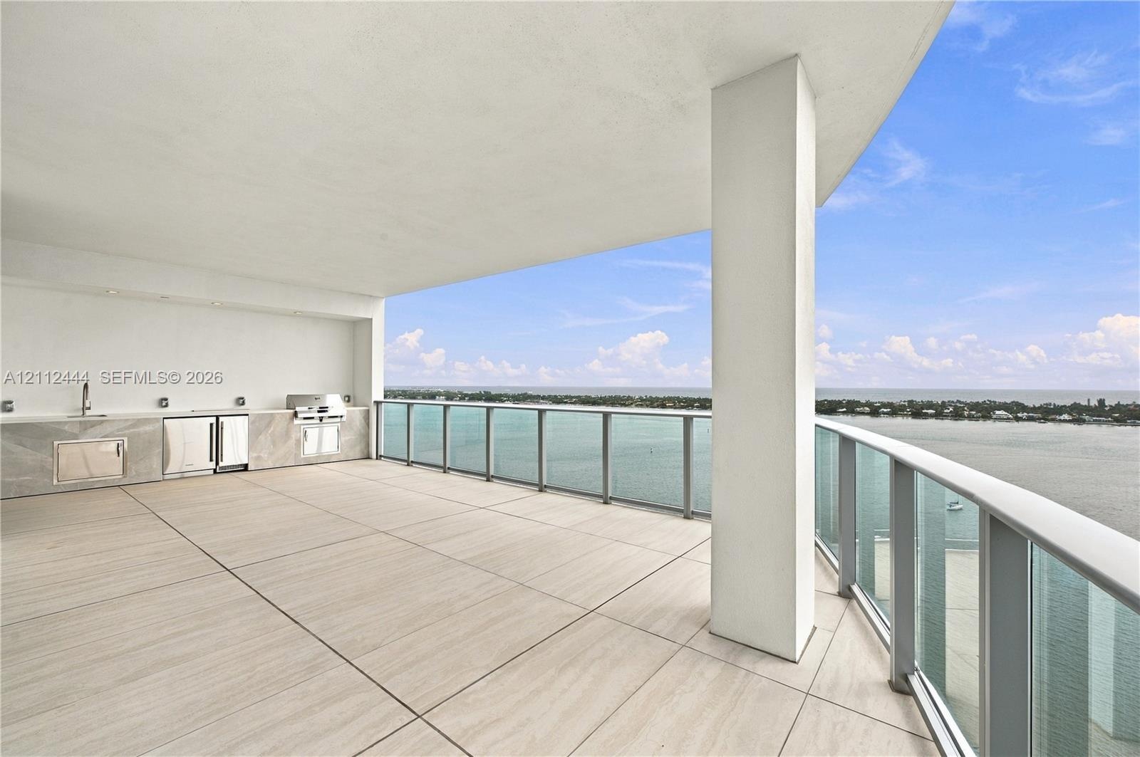 Covered terrace featuring large-format tile flooring, a built-in outdoor kitchen with stone-finish cabinetry, an integrated grill, and a glass-paneled