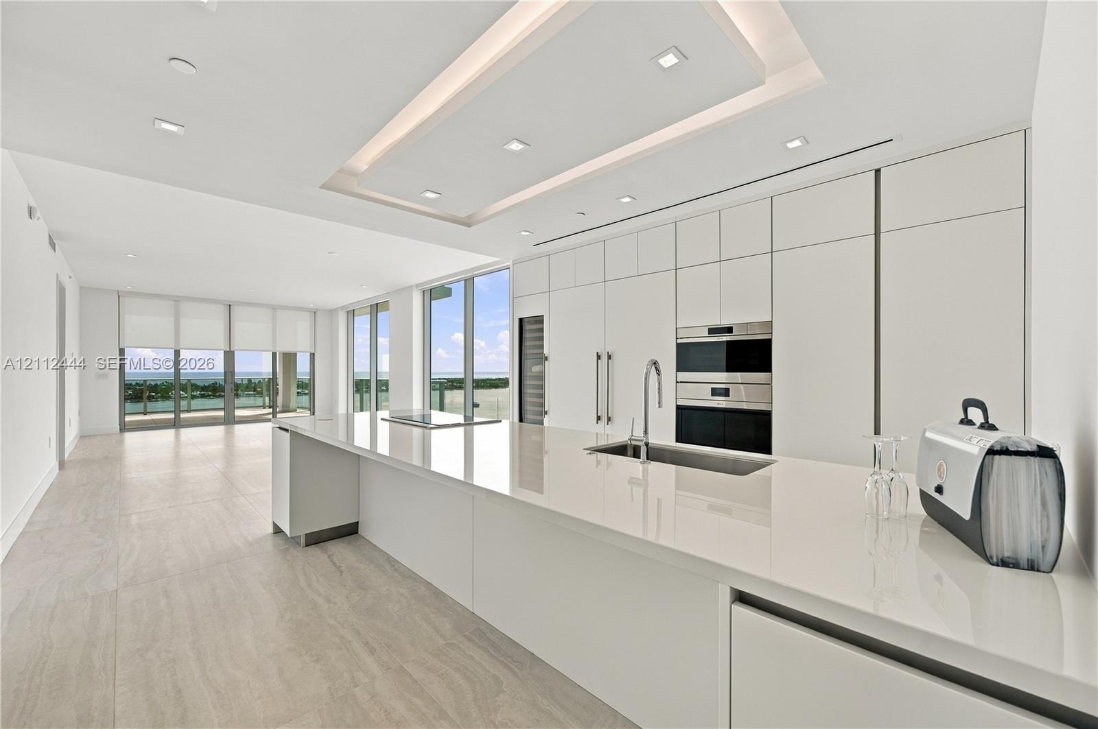 Modern kitchen featuring seamless flat-panel cabinetry, a spacious solid-surface island, built-in stainless steel appliances, recessed tray ceiling li
