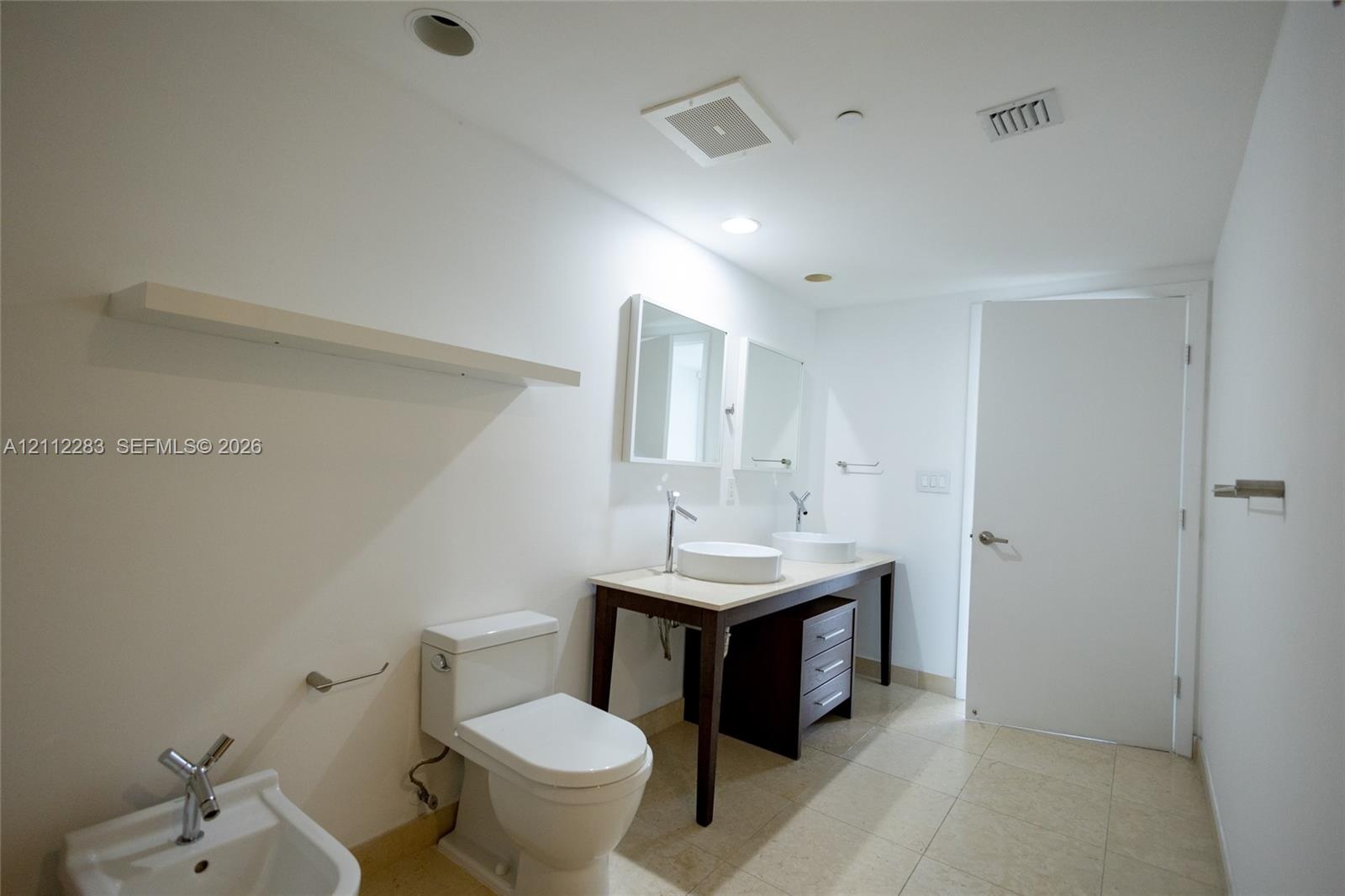 Dual-sink vanity with vessel basins, stone countertop, dark wood-finish cabinetry, ceramic bidet, and floating wall-mounted shelving