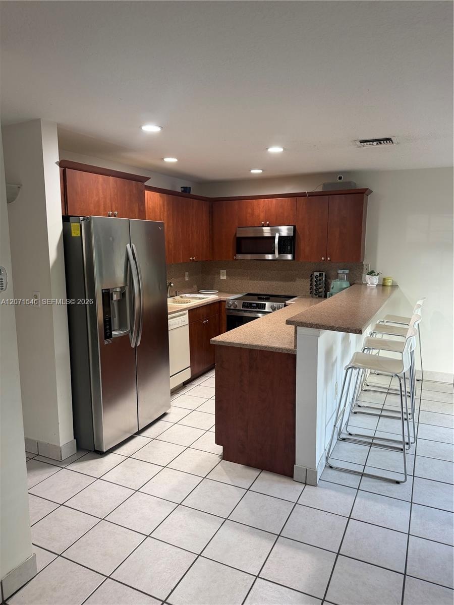 Kitchen featuring cherry-toned wood cabinetry, stainless steel appliances, solid-surface breakfast bar, and tile flooring
