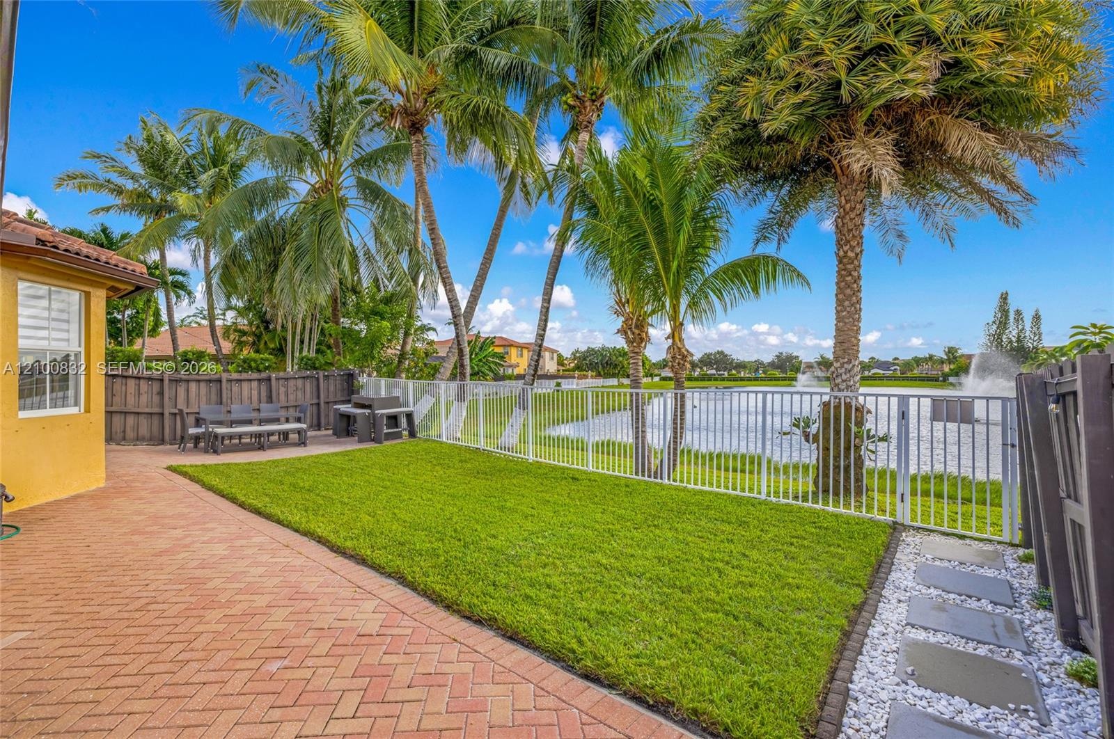 Fenced backyard featuring a brick-paver patio, manicured lawn, mature palm trees, and waterfront perimeter