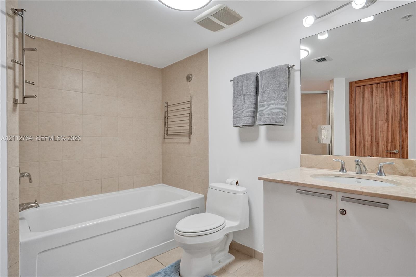 Bathroom featuring a deep soaking tub with neutral tile surround, vanity with stone-finish countertop, undermount sink, and full-width vanity mirror w