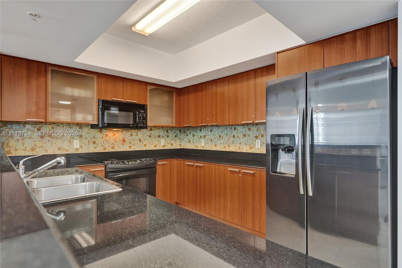 Kitchen featuring wood cabinetry, dark stone-finish countertops, multi-toned glass tile backsplash, recessed tray ceiling, and stainless steel applian