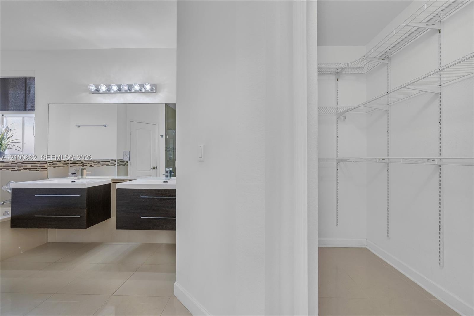 Double floating vanity with integrated rectangular sinks, oversized wall-to-wall mirror, multi-bulb vanity lighting, and walk-in closet featuring whit
