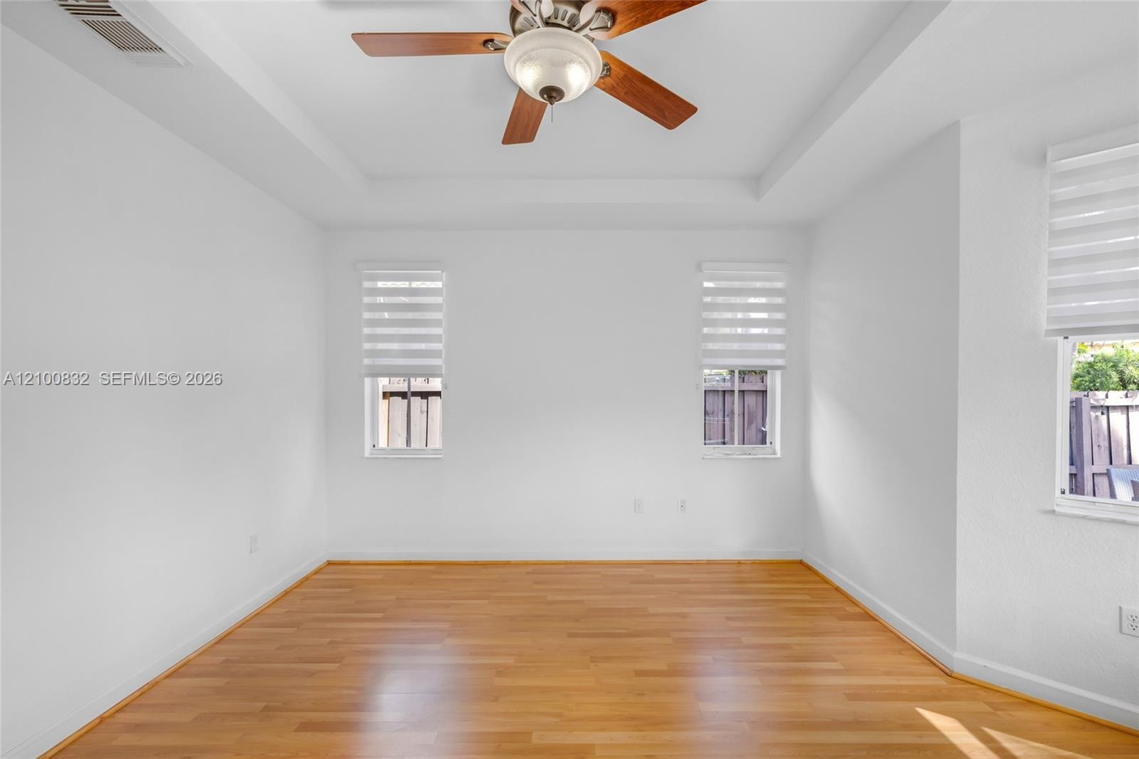 Room featuring a tray ceiling, wood-finish flooring, three windows with fitted horizontal blinds, and a central ceiling fan with integrated lighting
