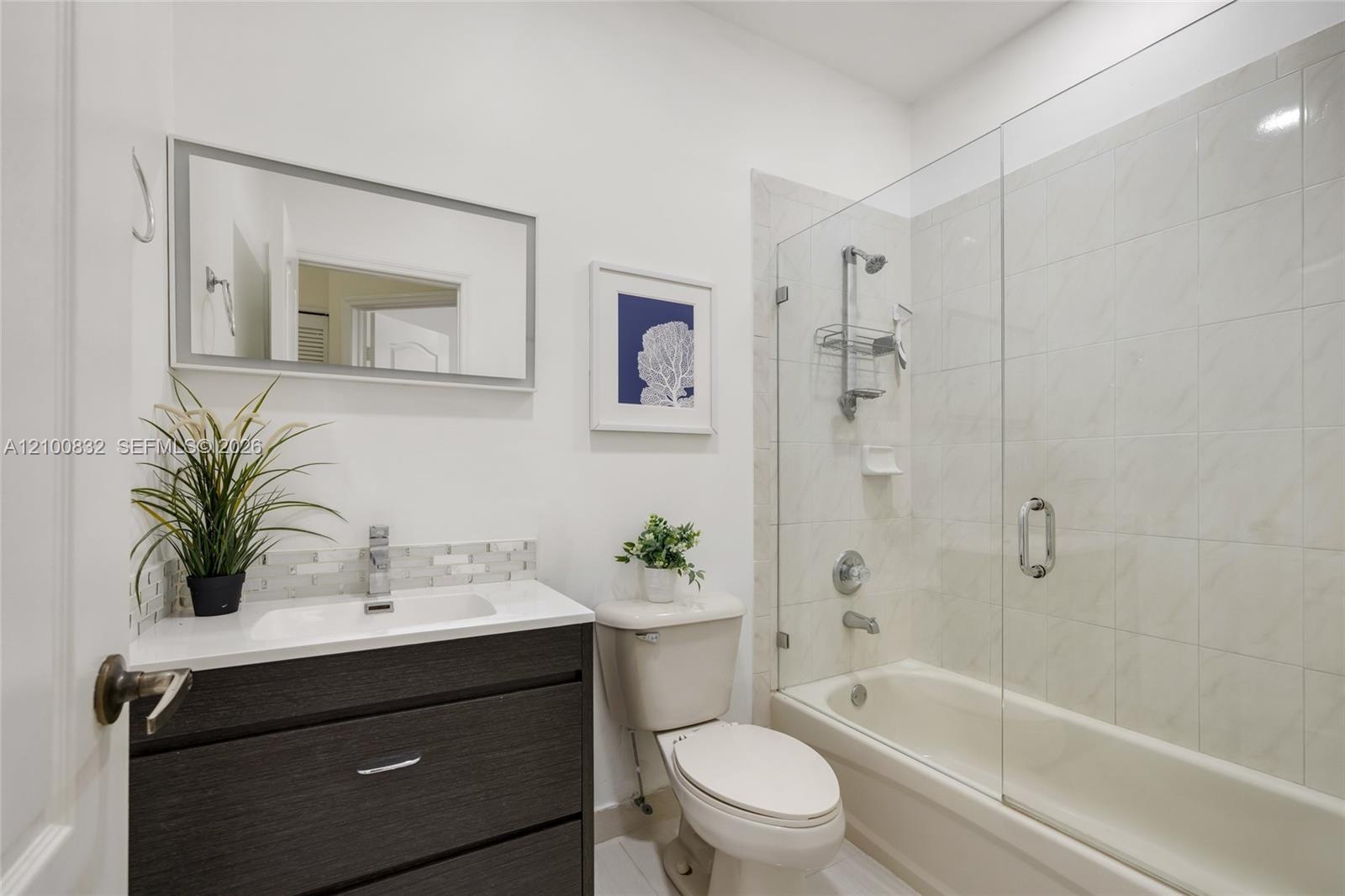 Bathroom featuring a dark wood-finish vanity with a white countertop, rectangular sink, glass-enclosed shower-tub combination, and tiled walls