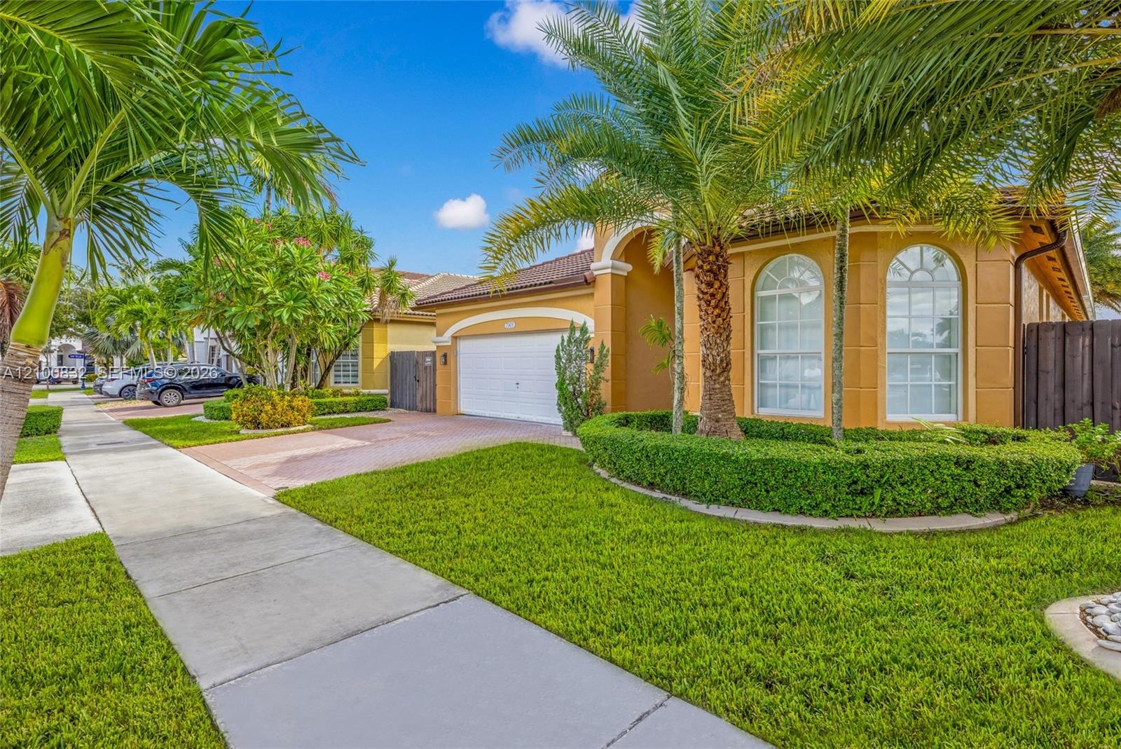 Stucco exterior featuring an arched entryway, paver driveway, two-car garage, and arched windows accented by landscaped shrubbery and palm trees