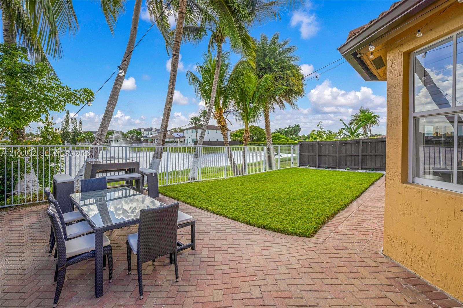 Outdoor space featuring brick paver patio, manicured lawn, white aluminum perimeter fencing, and mature palm trees