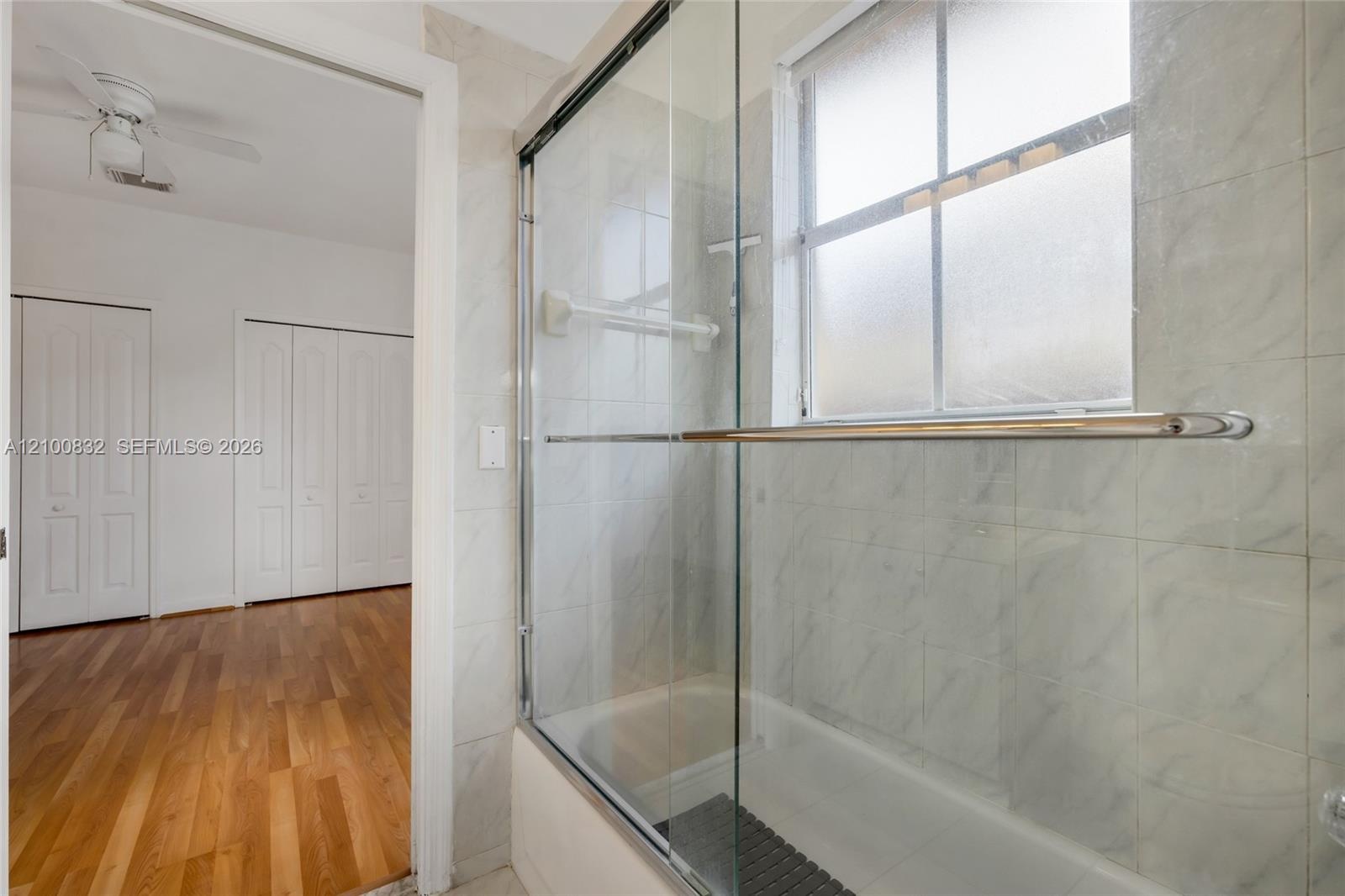 Bathroom featuring a glass-enclosed tub with metal hardware, light-toned wall tile, and a privacy-glass window