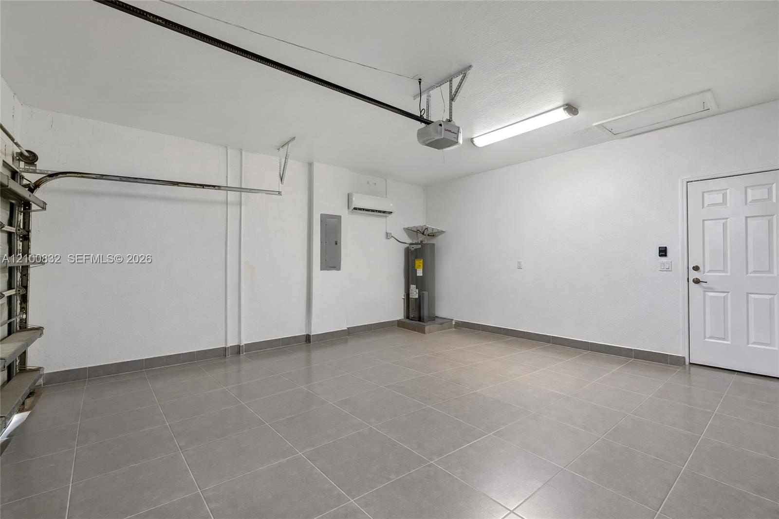 Garage featuring large-format tile flooring, white finished walls, wall-mounted climate control unit, water heater, and interior access door