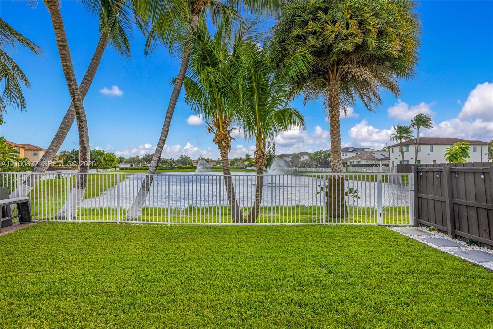 Lawn area featuring white metal perimeter fencing, mature palm trees, and a decorative water feature