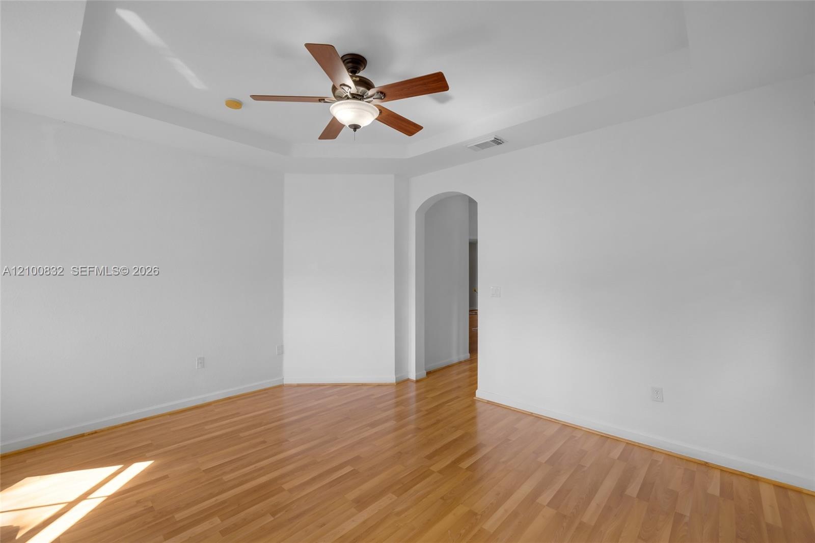 Tray ceiling with a central multi-blade ceiling fan, wood-finish flooring, and a decorative arched doorway