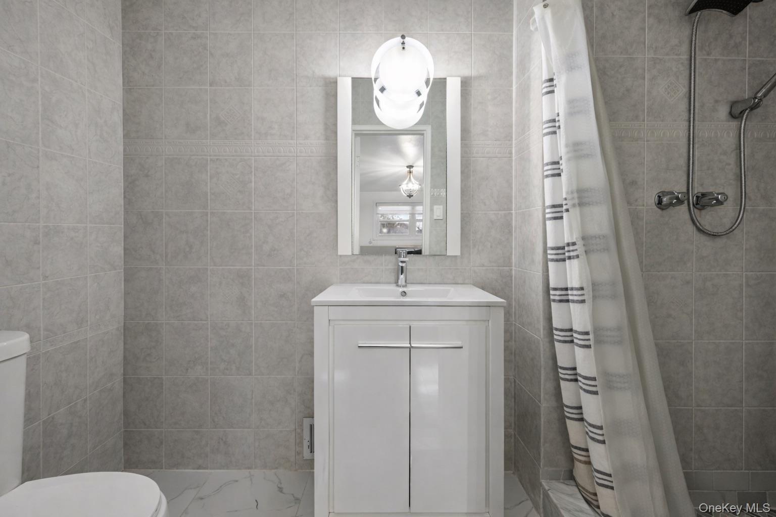 Bathroom featuring full-height neutral wall tiling, vanity with a white integrated sink, polished chrome faucet, wall-mounted globe lighting fixture, 