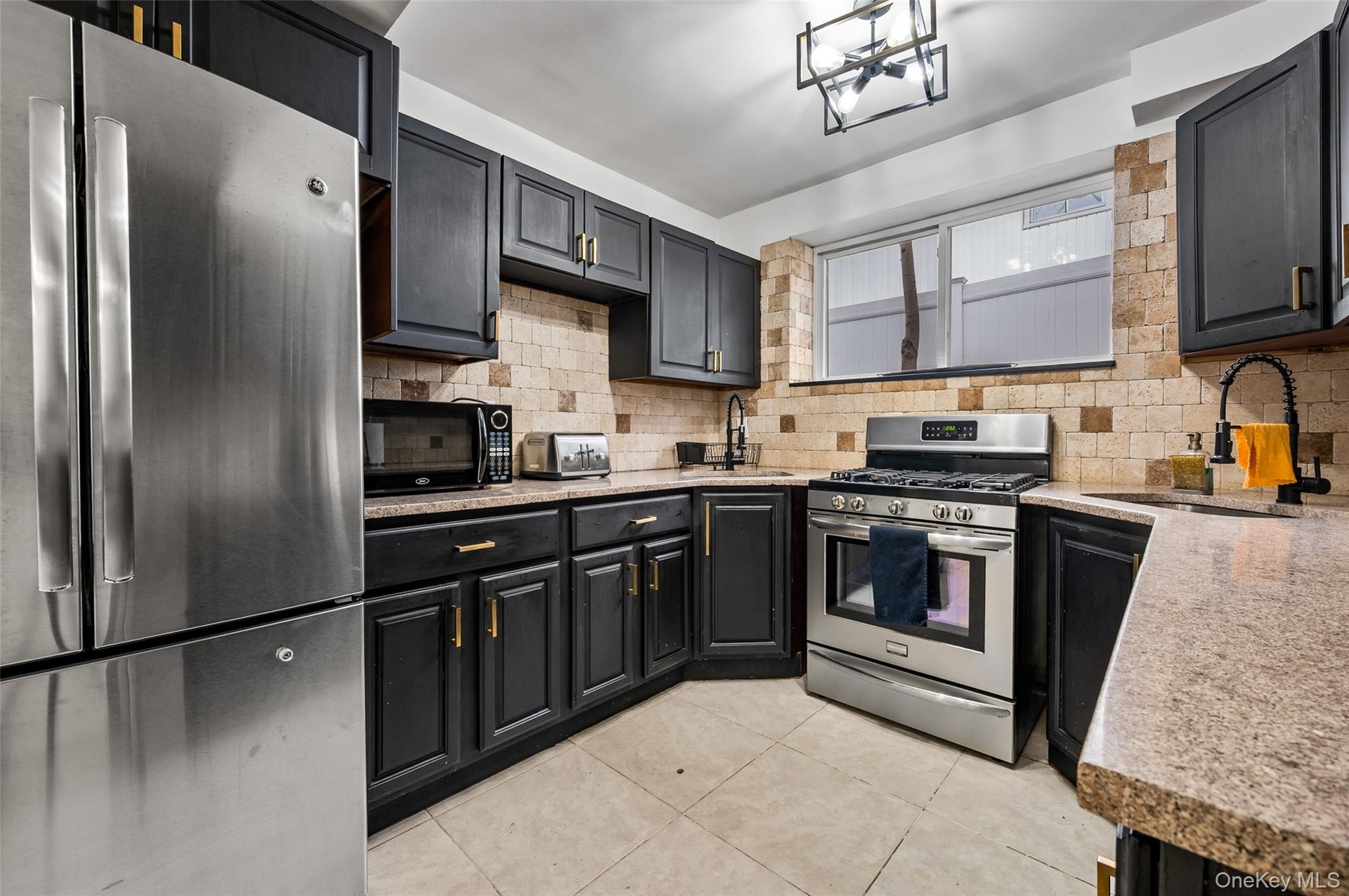 Kitchen featuring dark cabinetry with gold-tone hardware, stainless steel appliances, stone-surface countertops, stone tile backsplash, and tile floor