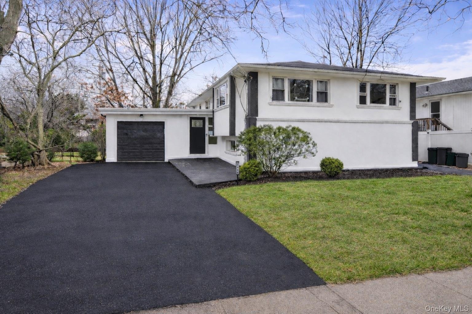 Split-level exterior featuring a light-toned smooth finish, dark-trimmed accents, an asphalt driveway, single-car garage, and landscape greenery