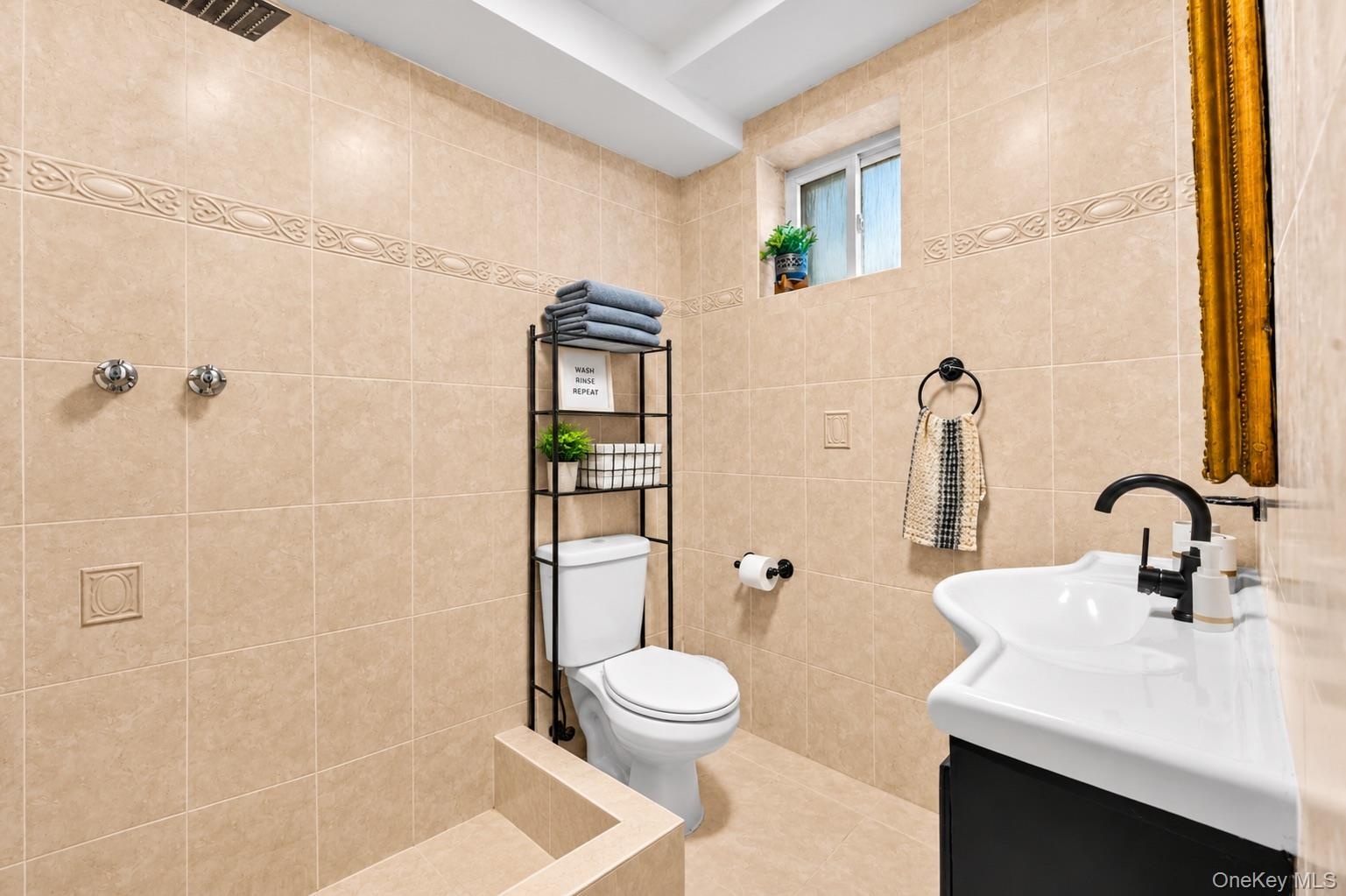 Full-wall neutral tile with decorative inlay, walk-in shower area, pedestal-style sink with matte black faucet, and recessed ceiling lighting