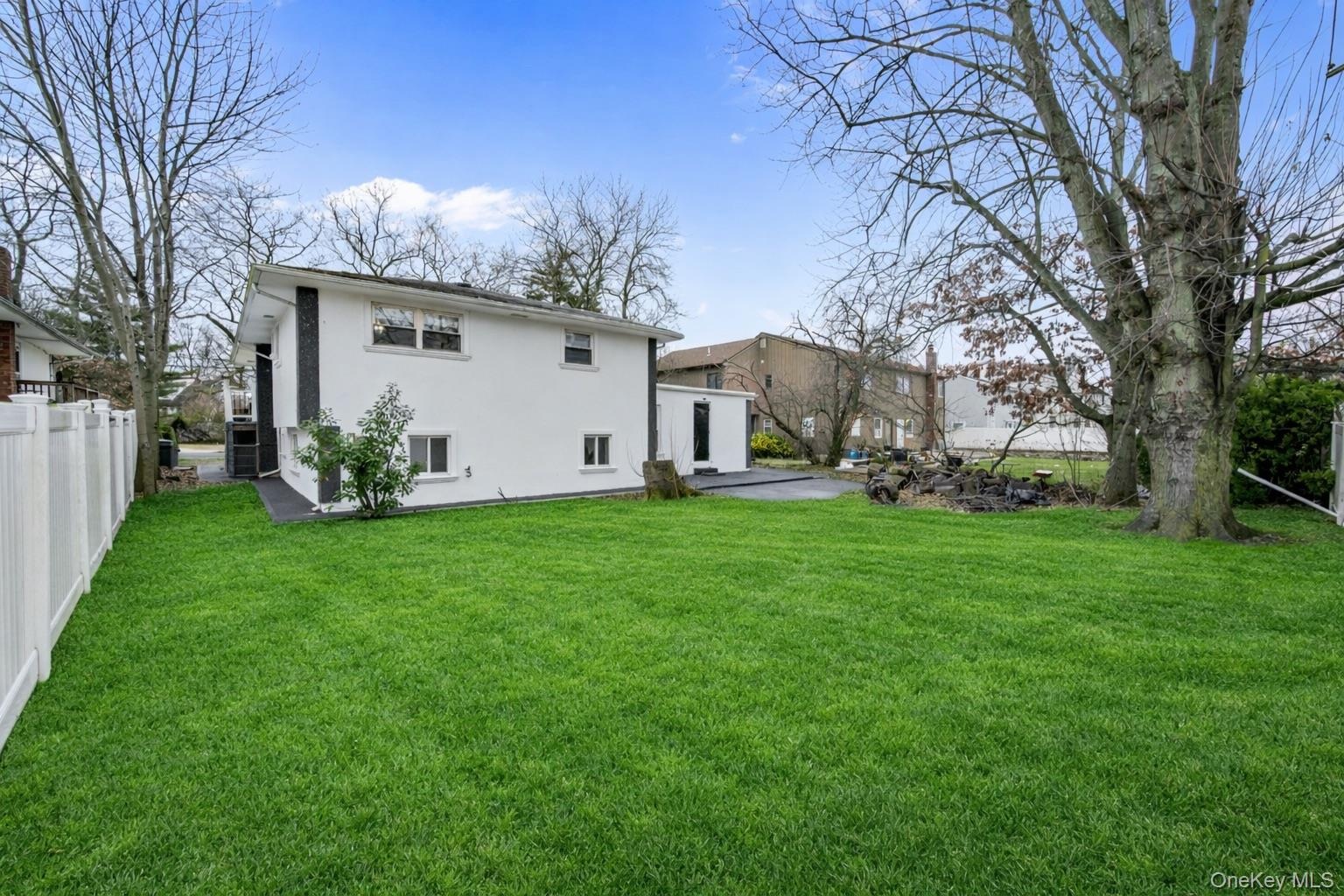 Expansive backyard featuring a level lawn, white vinyl fencing, mature trees, and a paved stone patio