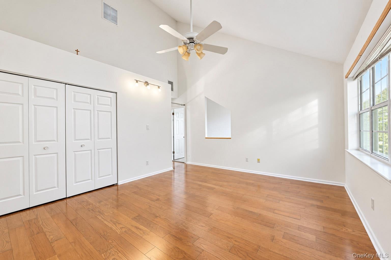 Vaulted ceiling featuring a central lighted ceiling fan, wood-finish flooring, multi-panel bi-fold closet doors, and large windows with natural light