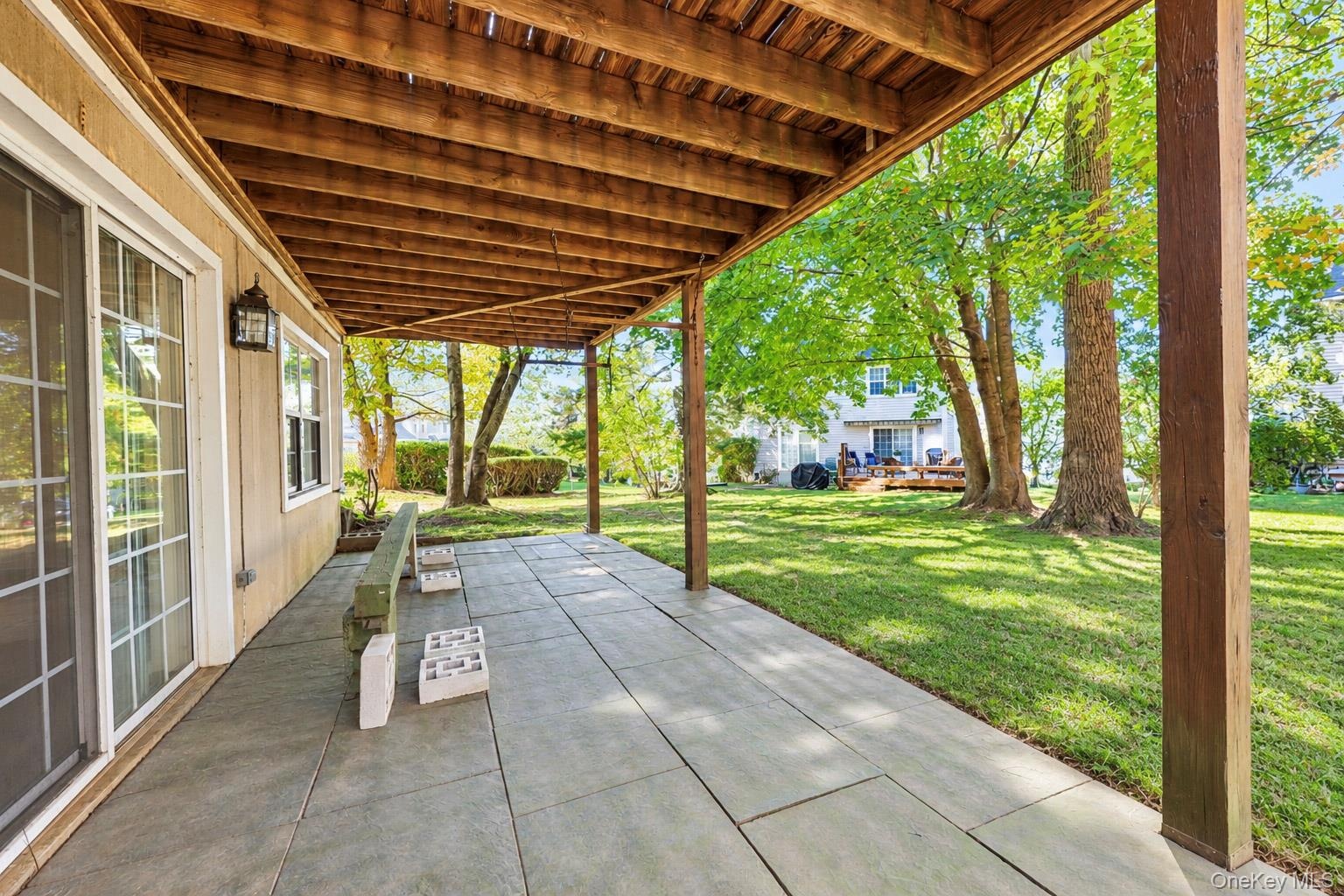 Covered patio featuring large-format stone-style pavers, exposed wood ceiling beams, sliding glass doors, and direct access to a lawn with mature tree