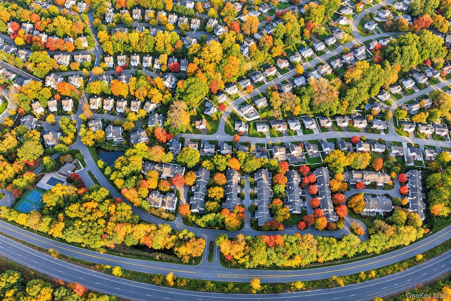 Aerial perspective of a residential community featuring mature deciduous trees, a tennis court, a swimming pool, and a winding internal road network