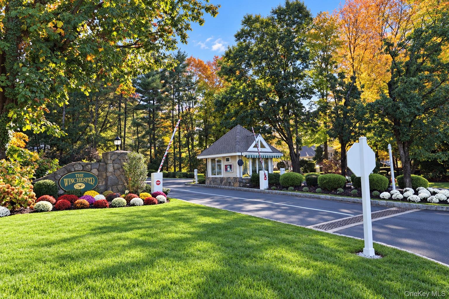 Gated entrance featuring a stone-clad guard house, asphalt driveway, automated security barrier, and professional landscaping