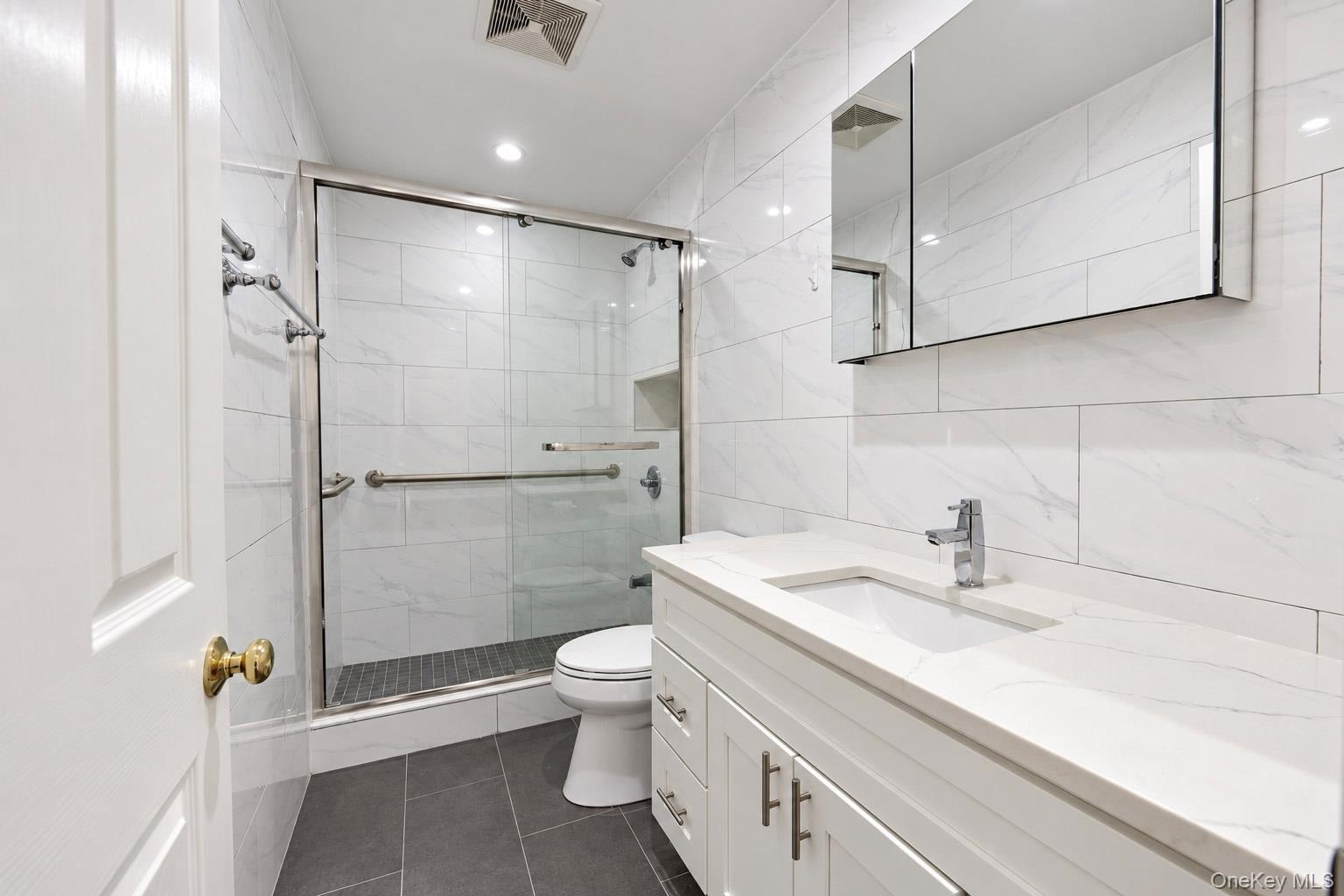 Bathroom featuring a white vanity with a composite countertop, undermount sink, mirrored medicine cabinet, glass-enclosed shower with tiled walls, and