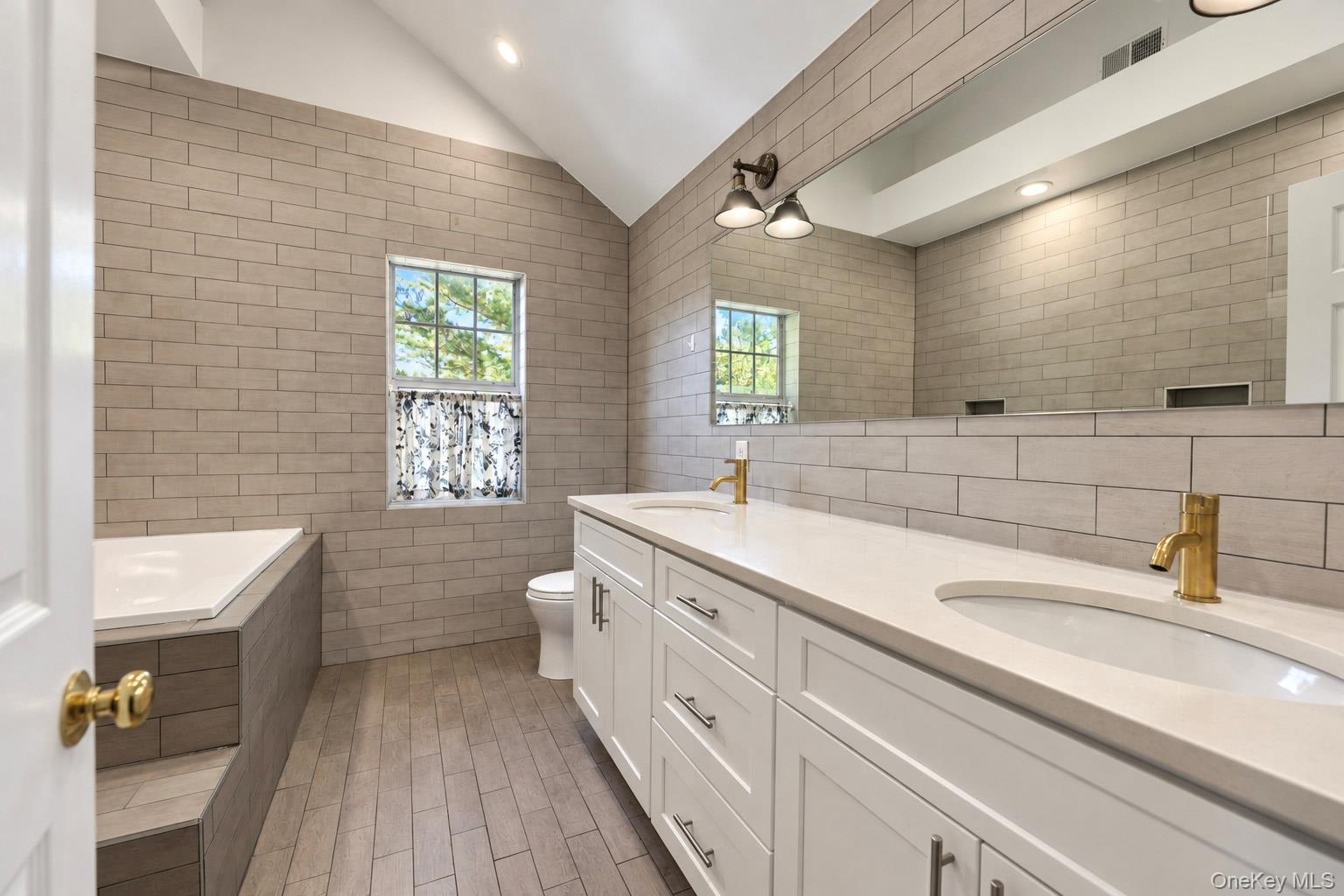 Bathroom featuring a dual-sink vanity with white shaker-style cabinetry, quartz countertops, and brass-finish fixtures, complemented by full-height ce