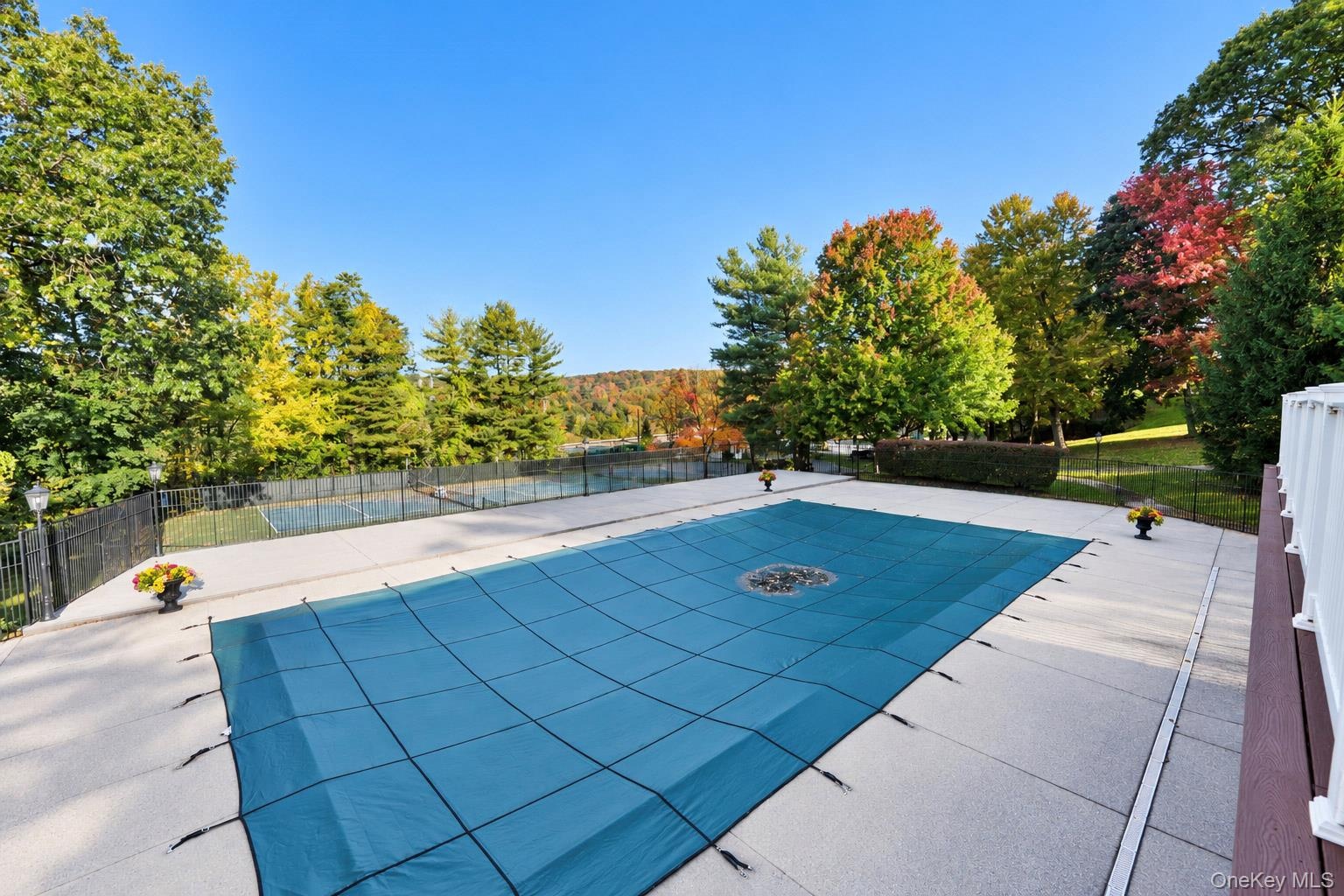 Outdoor area featuring a swimming pool with a mesh safety cover, expansive concrete pool deck, fenced tennis court, and mature trees