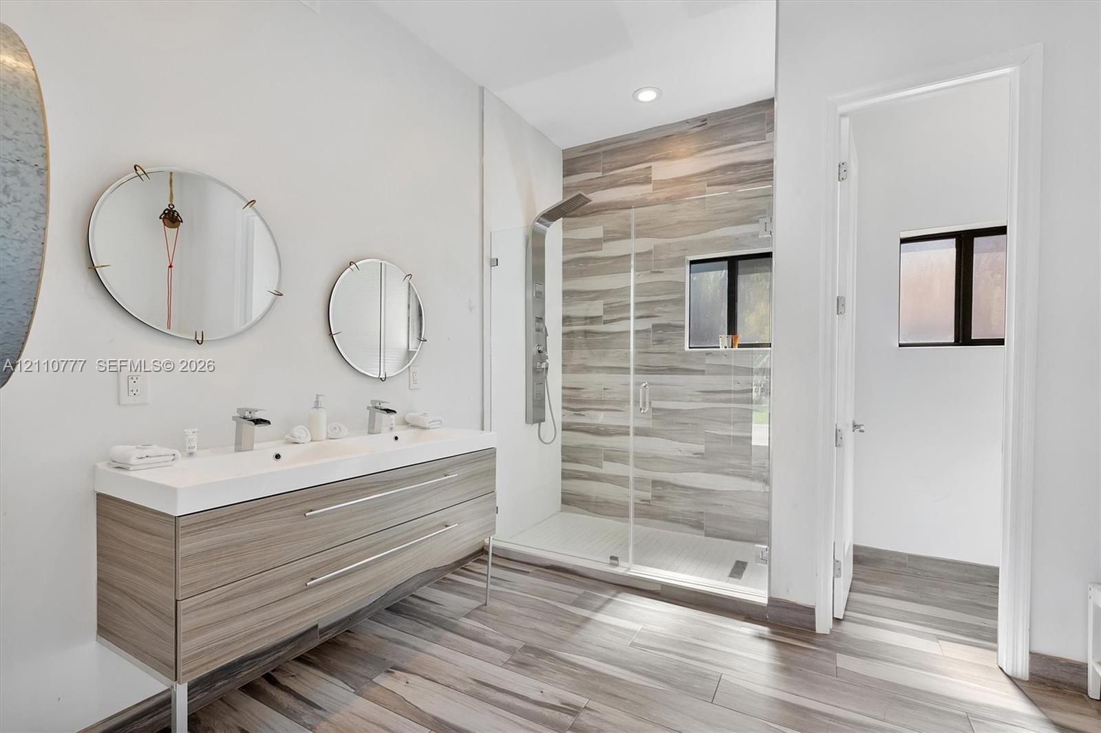 Double vanity with integrated sinks, wall-mounted faucets, wood-finish cabinetry, walk-in shower with horizontal tile surround, and wood-finish floori