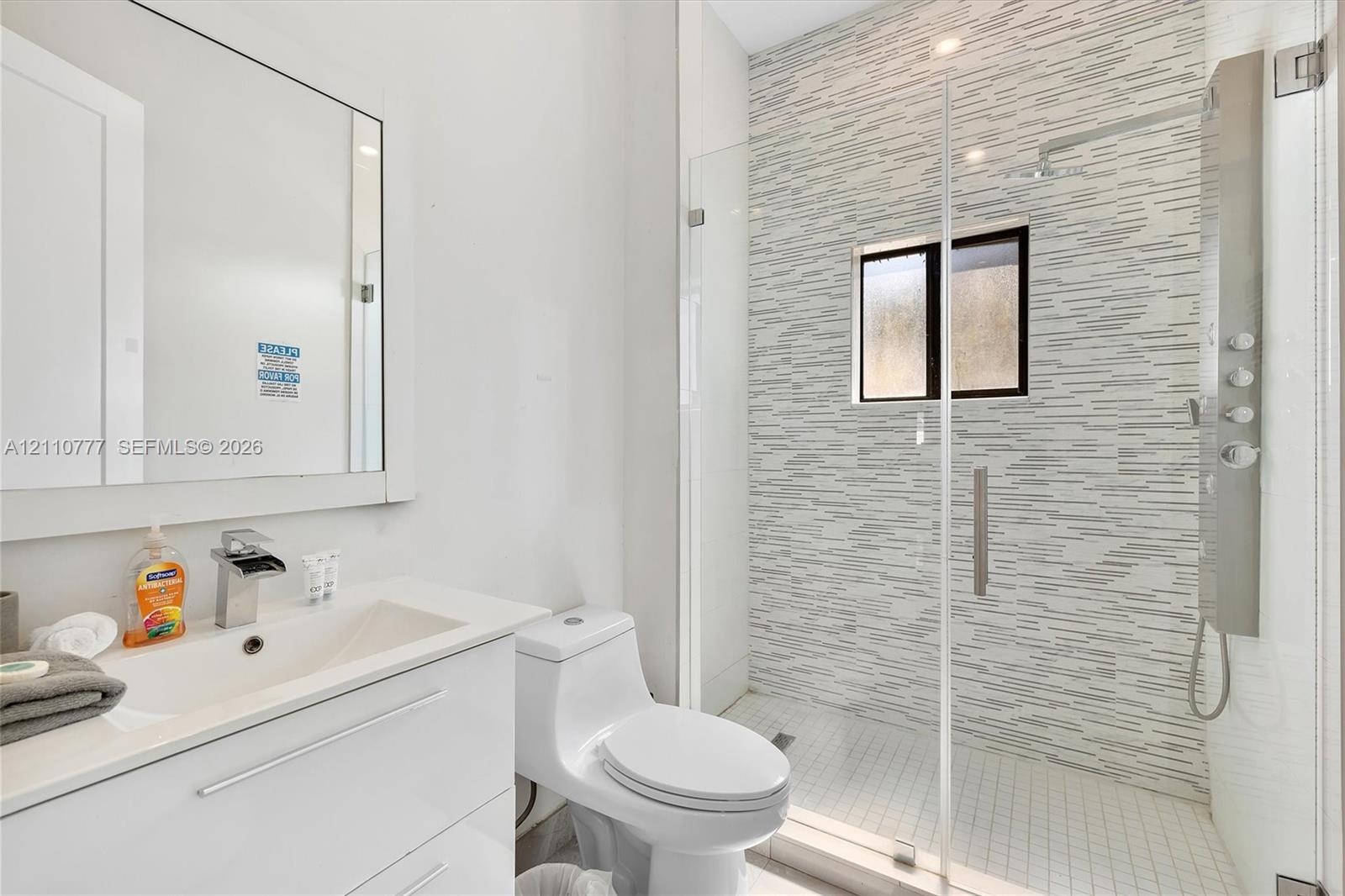 Modern bathroom featuring a white vanity with an integrated countertop, wall-mounted mirror, walk-in shower with a glass enclosure, patterned tile sho