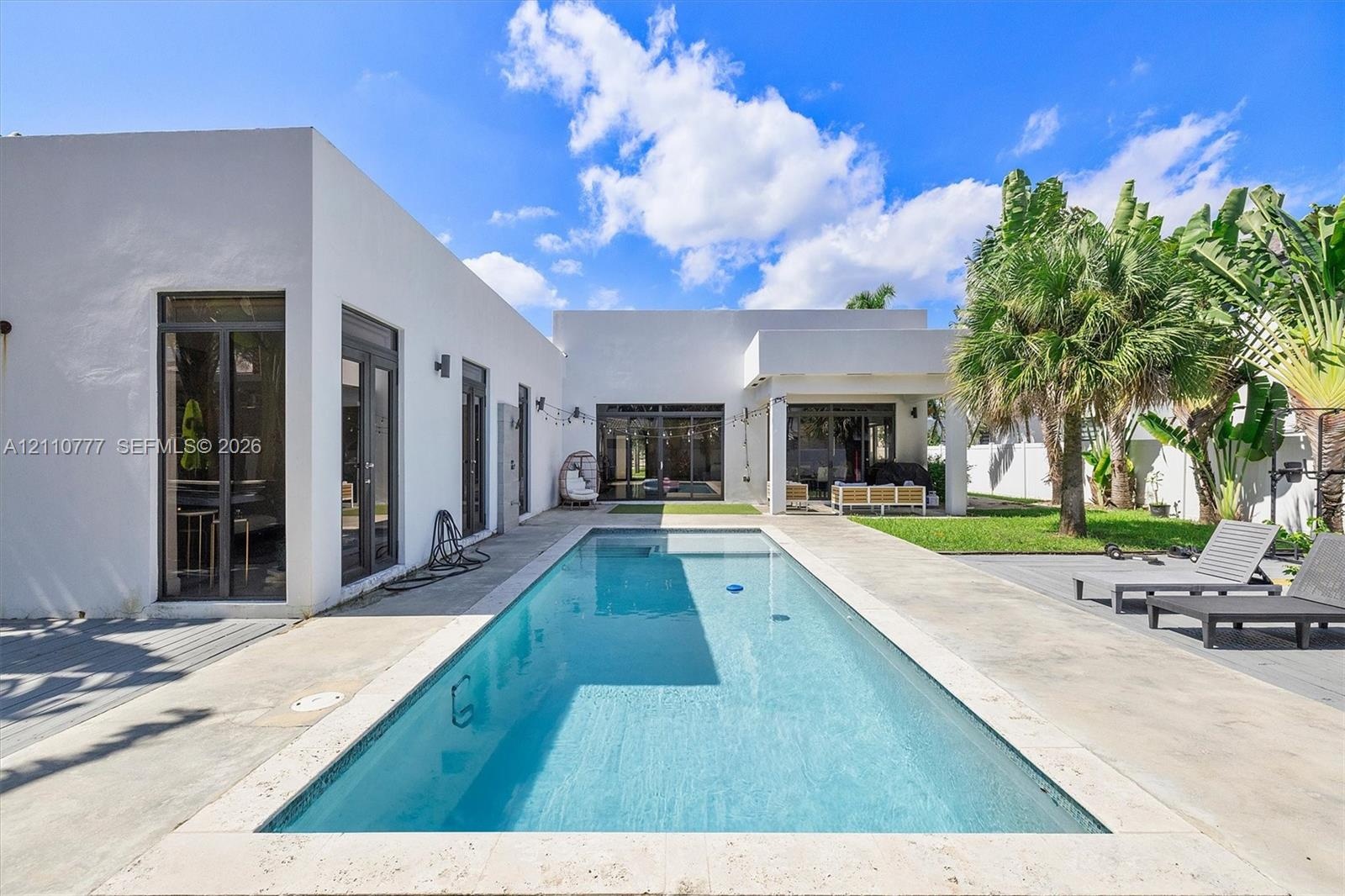 Modern exterior with smooth stucco finish, rectangular swimming pool with stone coping, concrete patio, covered outdoor area, and established palm tre