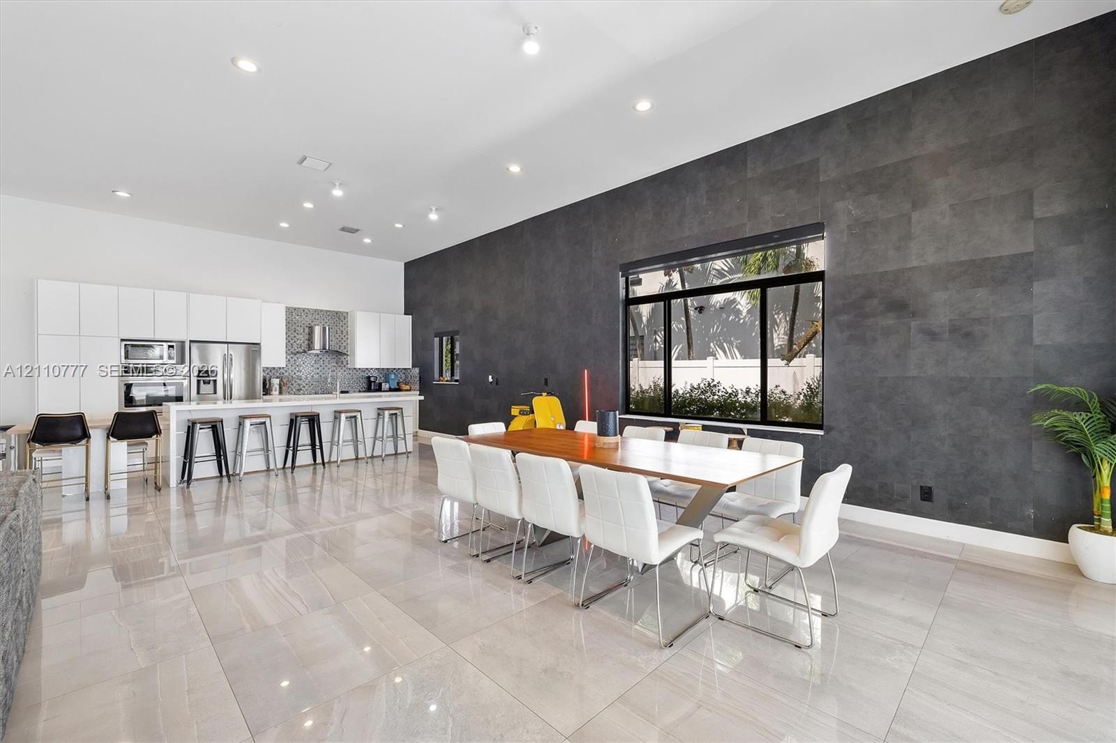 Modern kitchen and dining area featuring high-gloss white cabinetry, expansive stone tile flooring, a dark charcoal textured accent wall, recessed lig