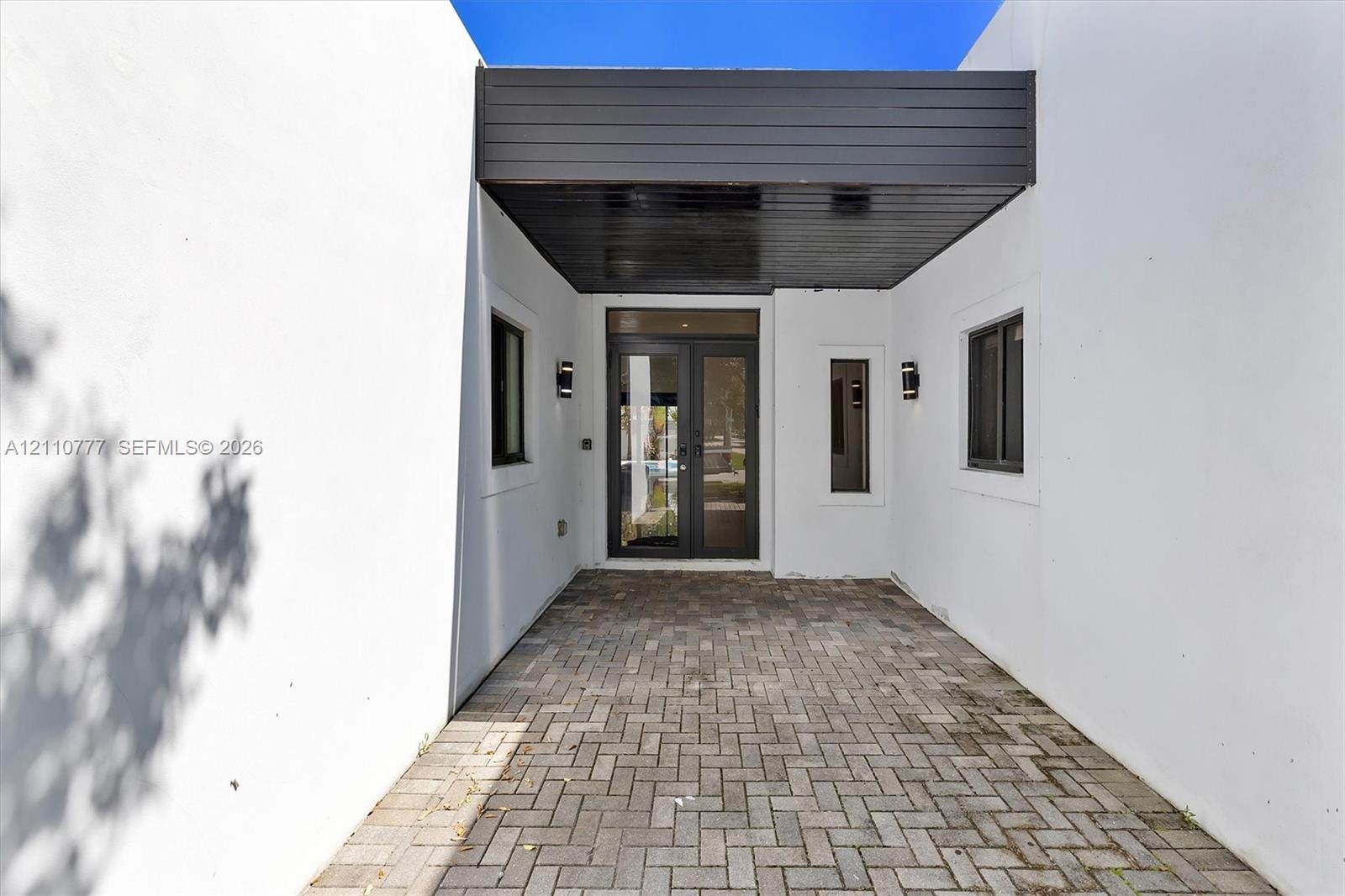 Covered entryway featuring a herringbone-patterned paver floor, minimalist smooth-stucco exterior walls, dark wood-finish ceiling paneling, glass-pane