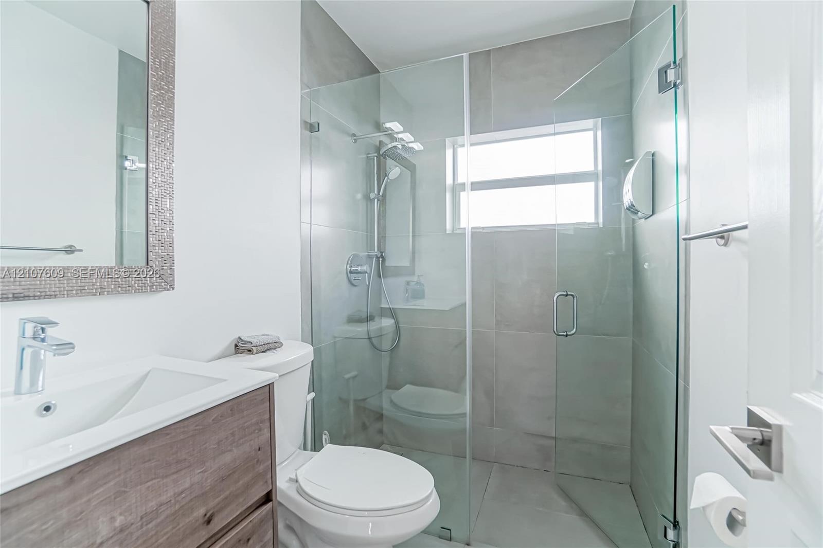 Bathroom featuring a wood-finish vanity with a rectangular basin, glass-enclosed shower with large-format tile surround, integrated shower head system