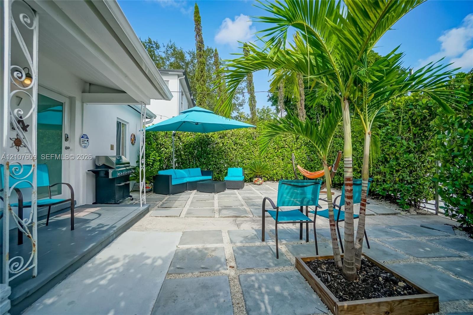 Rear patio featuring concrete paver hardscaping, white exterior walls, decorative metal porch pillars, and a dedicated garden bed with a mature palm