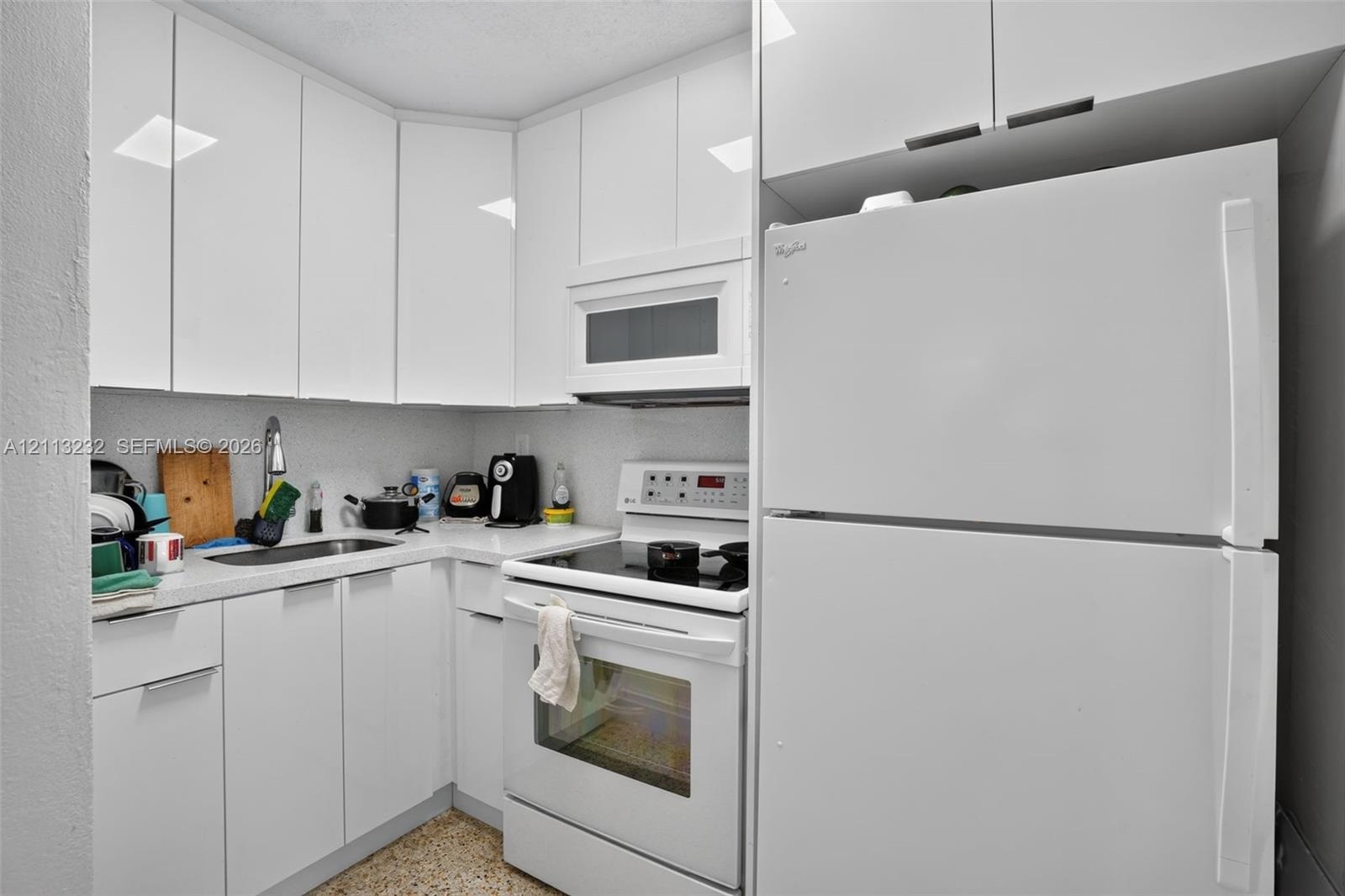 Kitchen featuring high-gloss white cabinetry, solid-surface countertops, stainless steel sink with high-arc faucet, and white appliances including a b